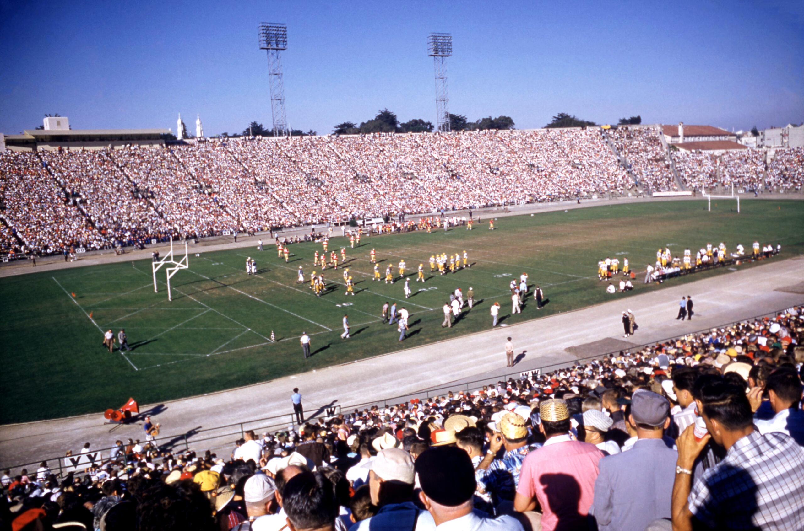 A crowded stadium with spectators watches a football game. Players in uniforms are on the field, with coaches and staff nearby. The sky is clear and blue.