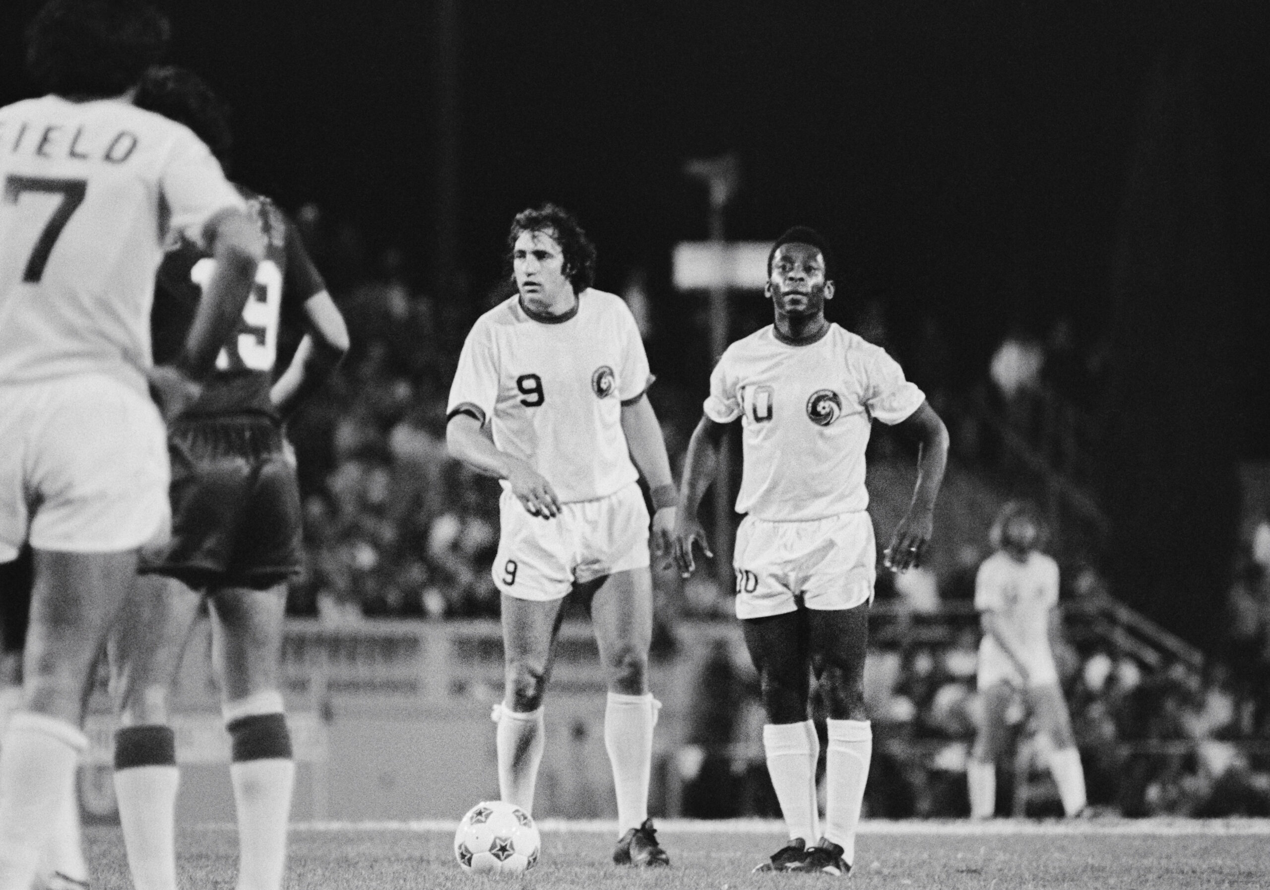 Two soccer players stand on a field, wearing jerseys with numbers 9 and 10. They are focused, with others visible in the background and near the ball.