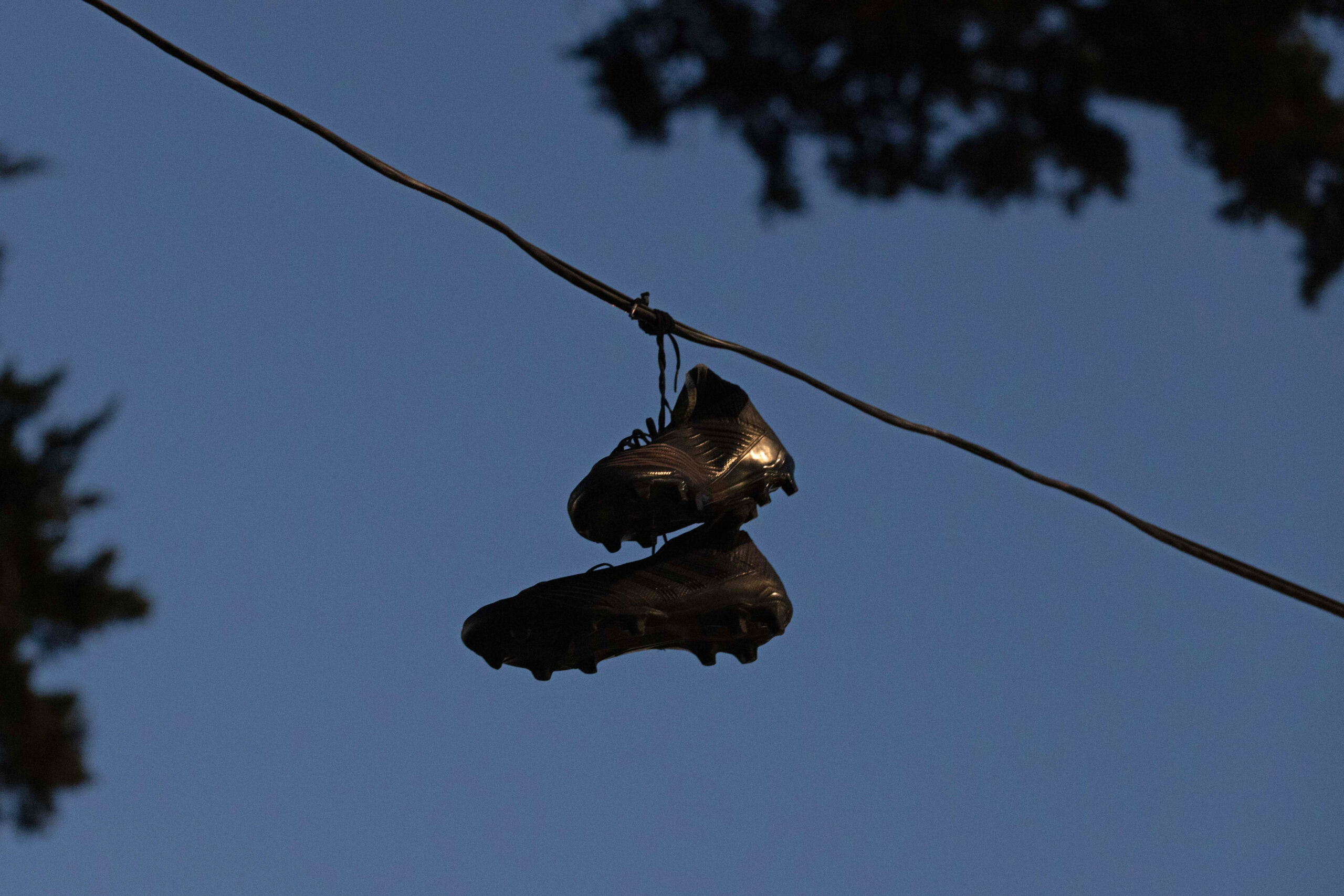 A pair of black soccer cleats is hanging by their laces over a wire, set against a clear blue sky, with partial silhouettes of trees in the background.