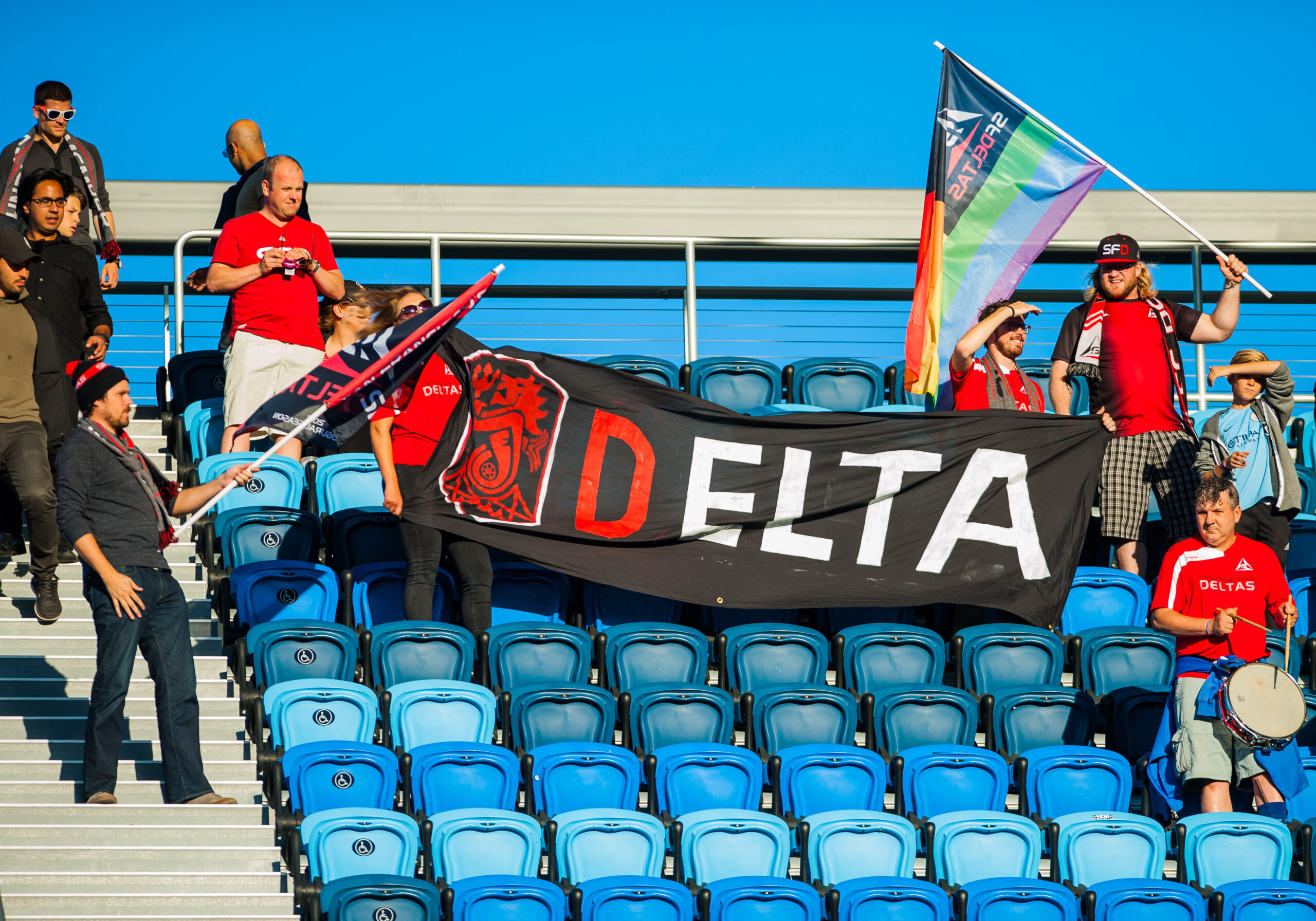 A small group of people, mostly in red shirts, stand in nearly empty blue stadium seats. They hold a large &quot;DELTA&quot; banner and a rainbow flag.