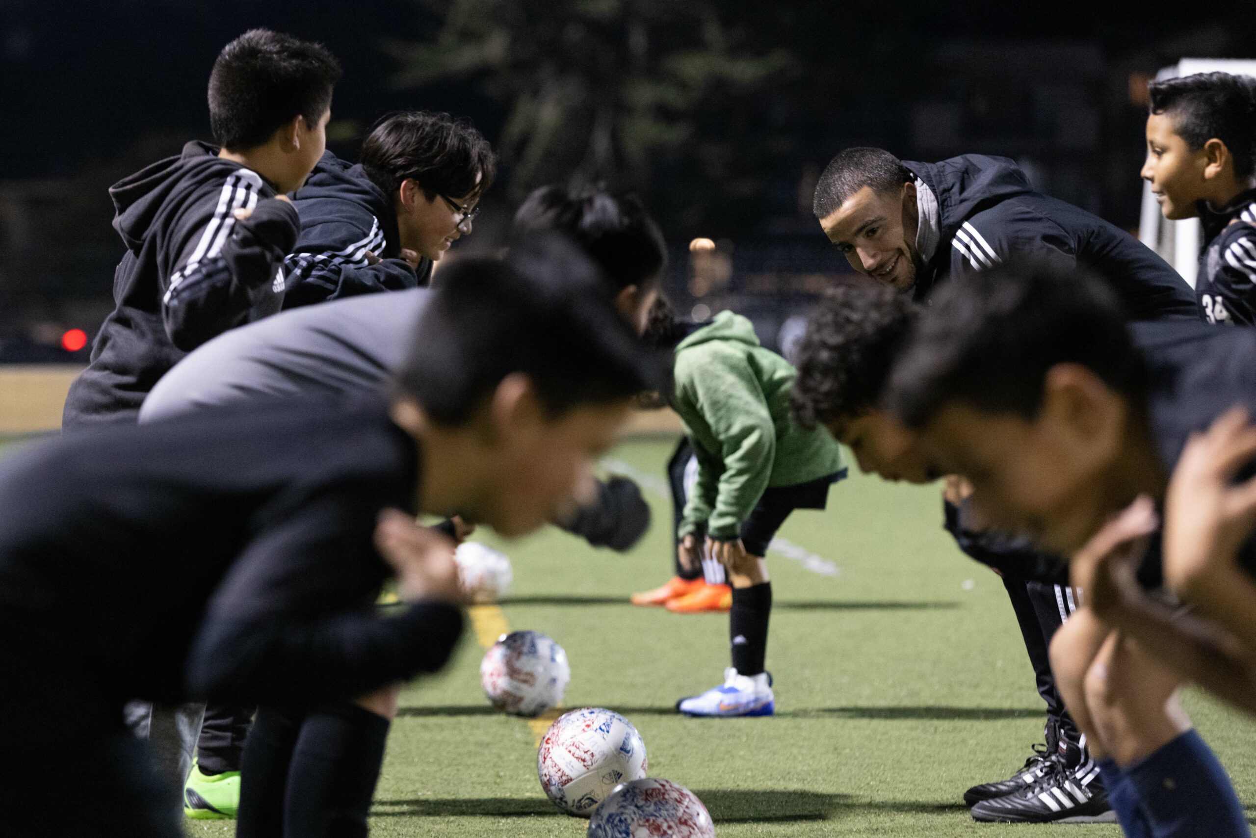A group of young soccer players and their coach are bent over in a line on a grass field at night. They are dressed in sports clothing, smiling and engaged.