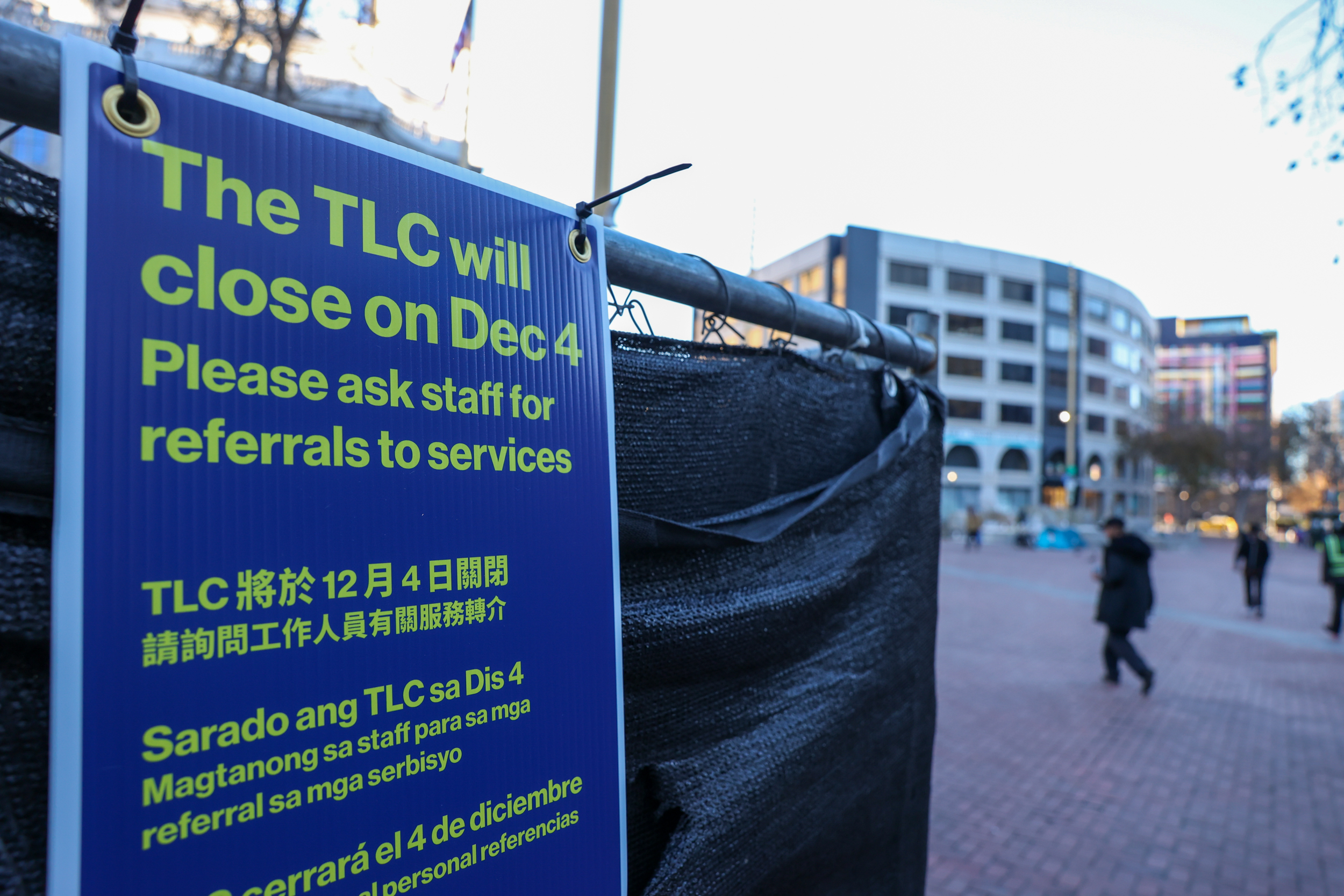 A sign stating "The TLC will close on Dec 4" in multiple languages, attached to a fence, with an urban background.