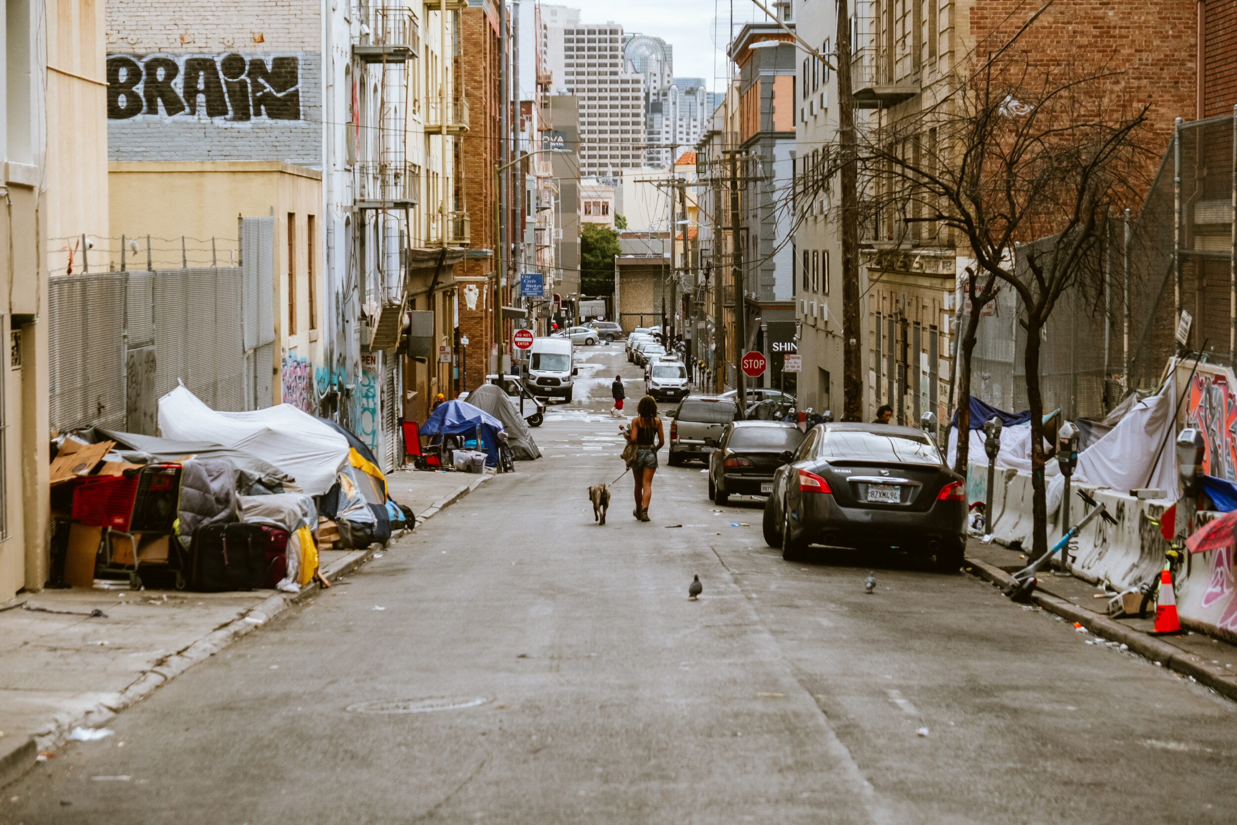 A city alley with tents, debris, a person walking a dog, cars, and distant buildings.