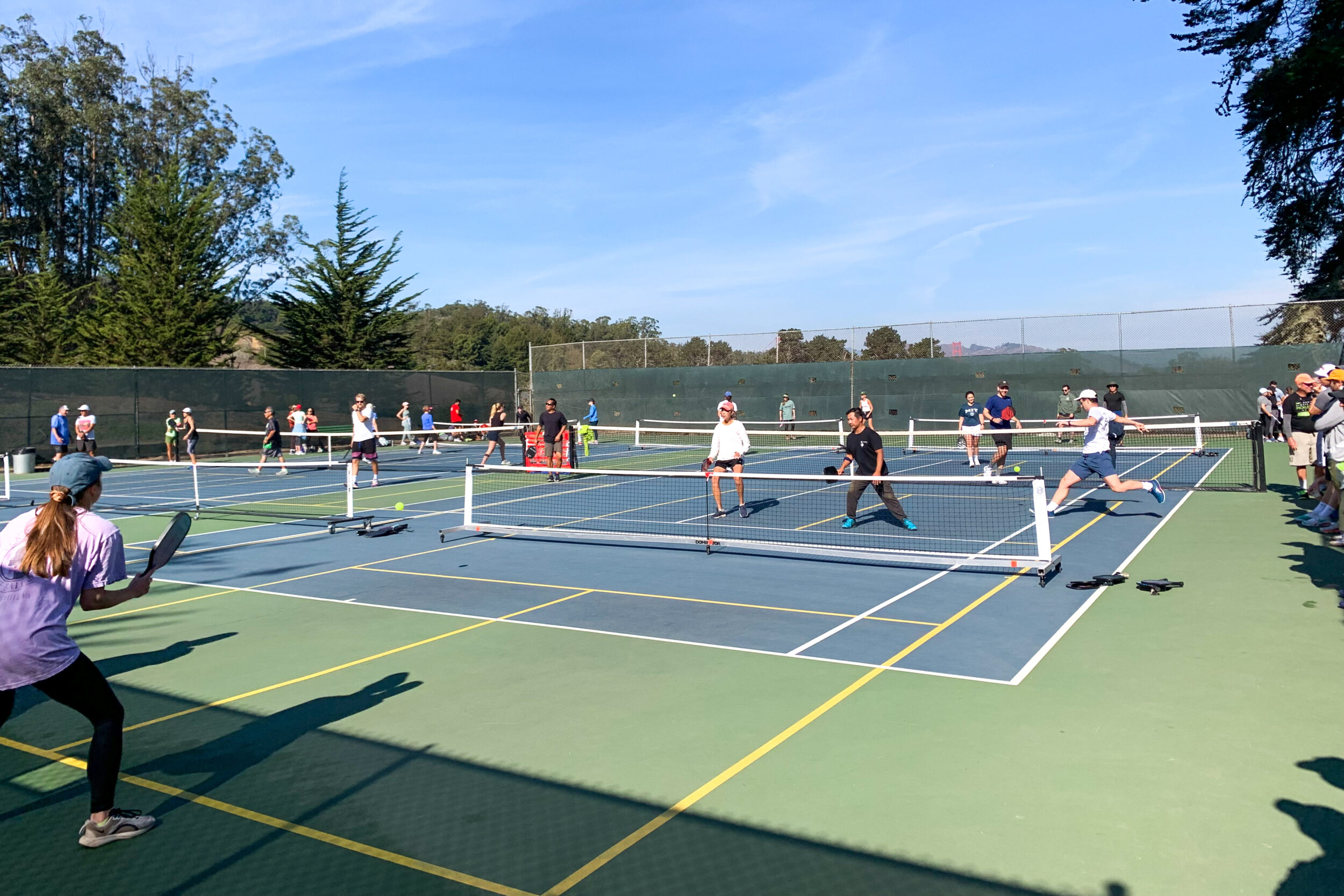 This image shows multiple people playing pickleball on outdoor courts on a sunny day.