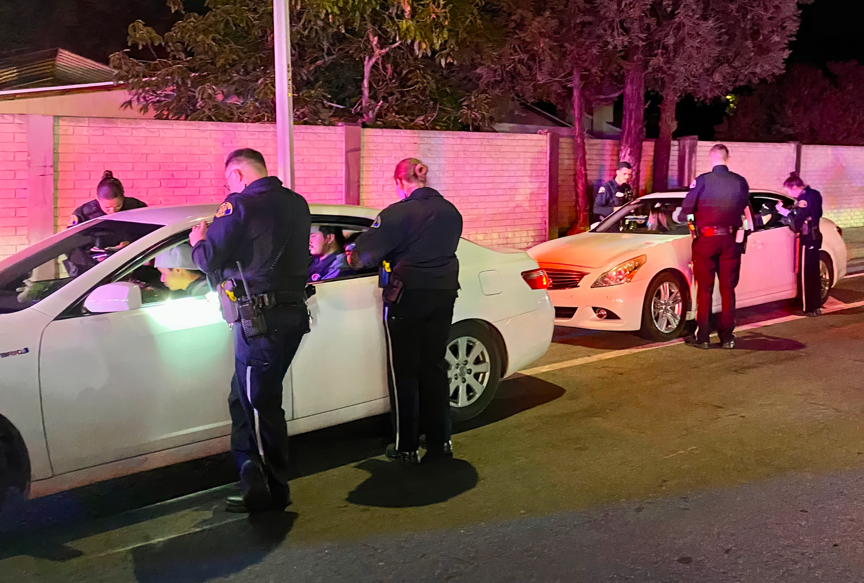 Two police officers in uniform shine a flashlight into a parked white sedan parked next to a large wall at nighttime.