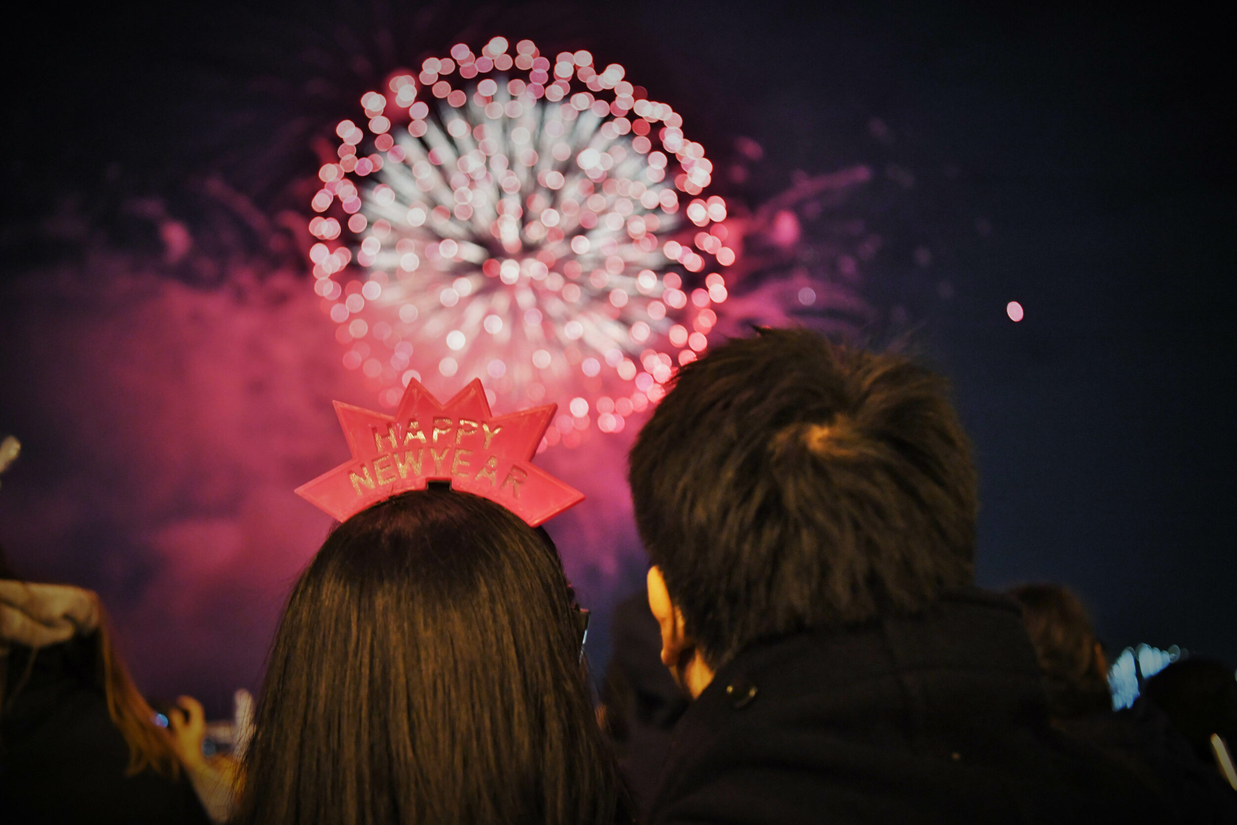 The back of two people's heads with brown hair watch a red and pink fireworks display.