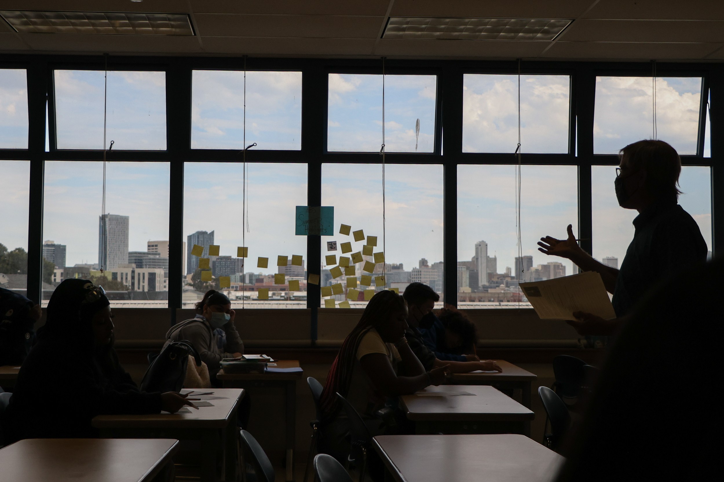 A classroom setting with students at desks, a person speaking in front, and a window view of a city skyline. Sticky notes are on the window.