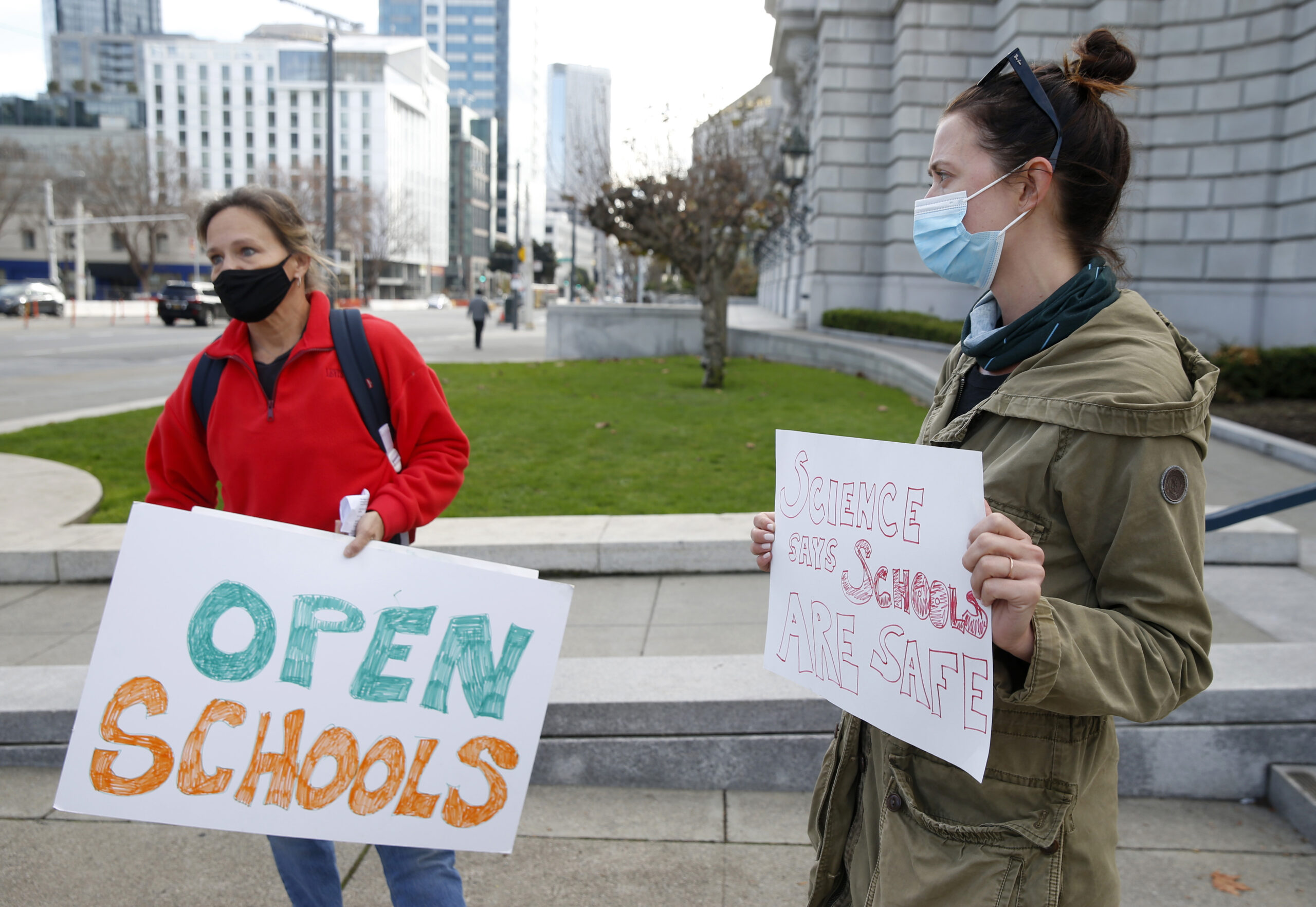 Two masked individuals stand outside holding signs advocating for opening schools, with buildings and a lawn in the background.