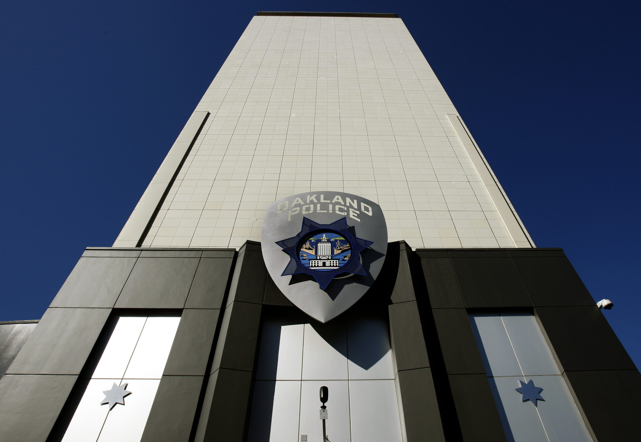 A multi-story administration building with an Oakland police shield affixed to its exterior stands against dark blue skies in direct sunlight.