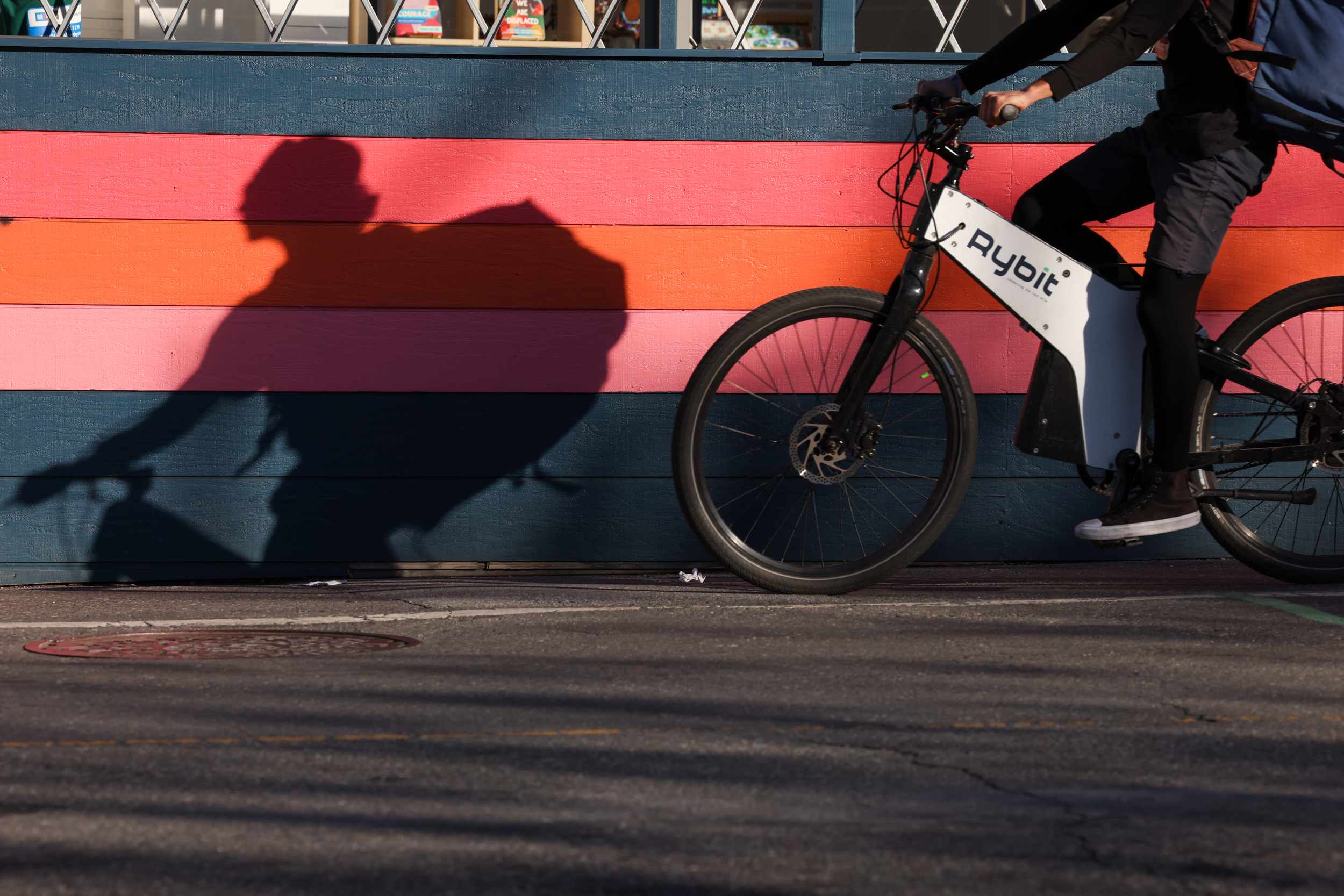 A cyclist casts a sharp shadow on a colorful, striped wall while riding an e-bike.