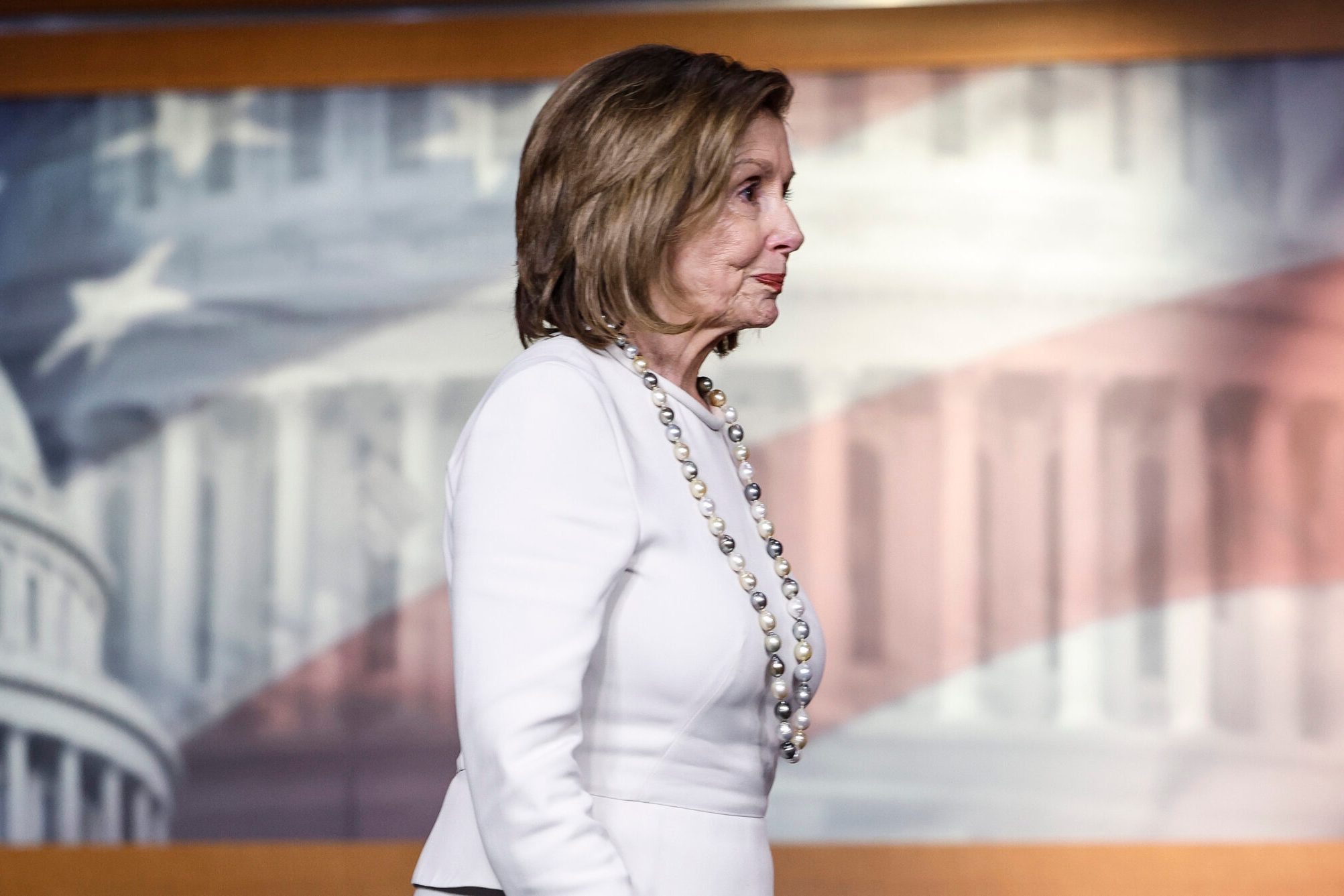 A woman in a white suit and pearl necklace stands sideways in front of a backdrop featuring a blurry image of the U.S. Capitol Building and American flag.