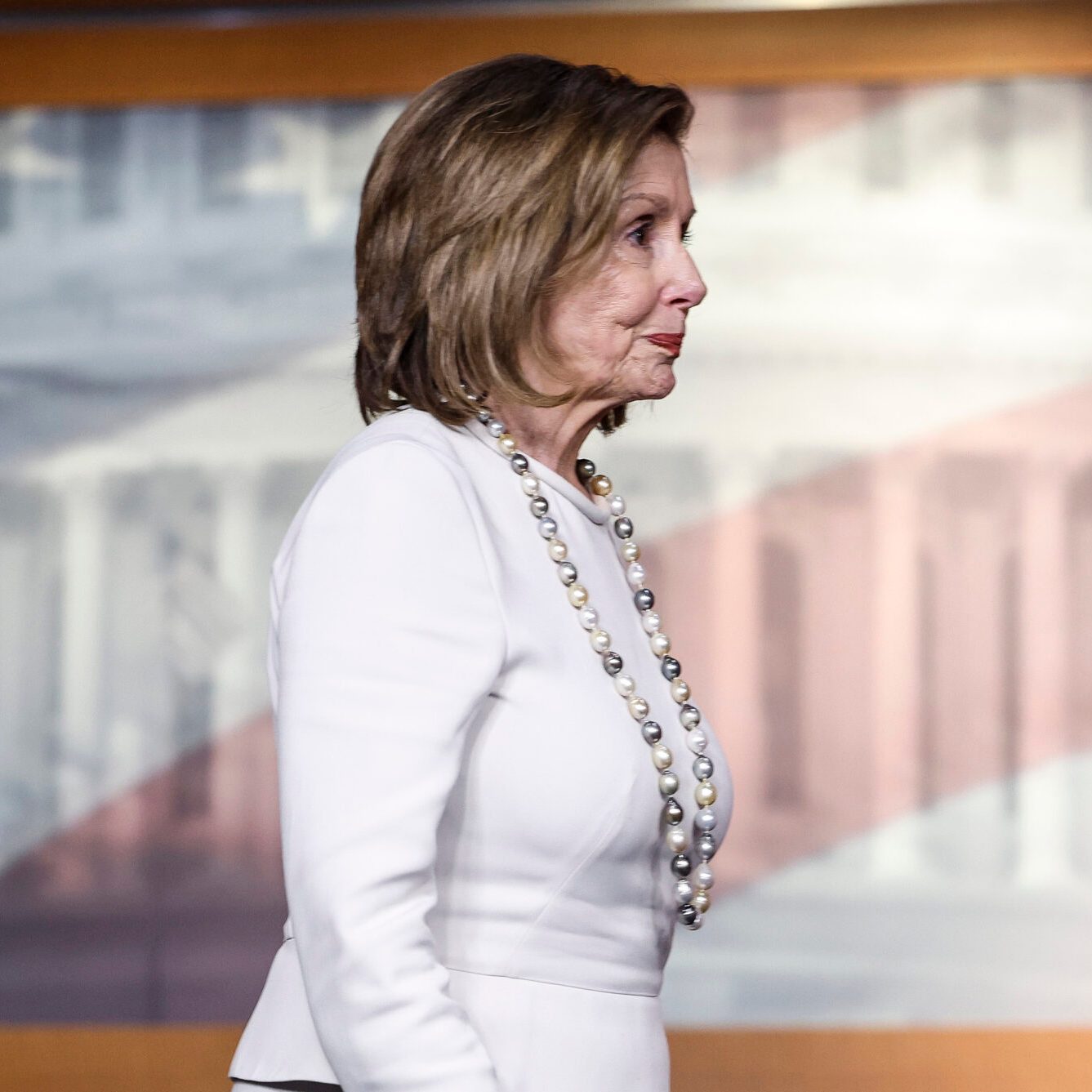 A woman in a white suit and pearl necklace stands sideways in front of a backdrop featuring a blurry image of the U.S. Capitol Building and American flag.