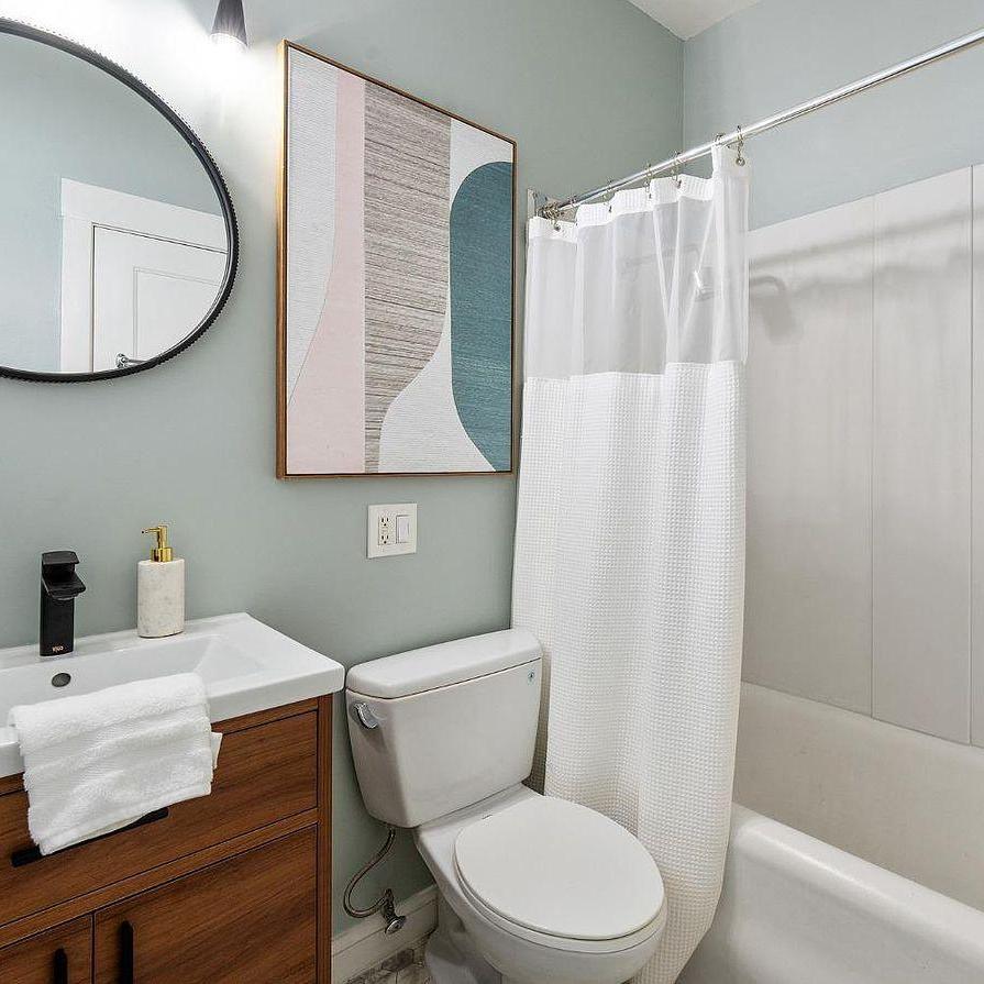 The image shows a modern bathroom with a bathtub and white shower curtain, a toilet, and a wooden vanity with a round mirror above it.