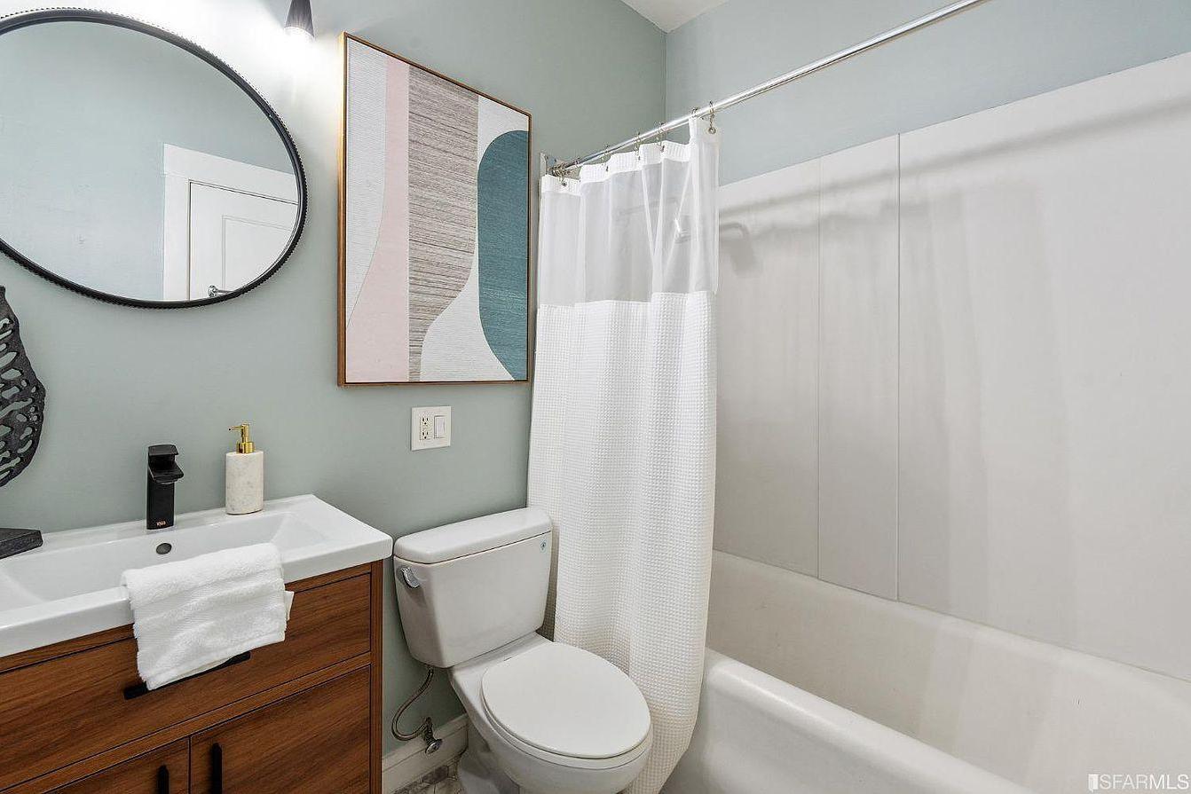 The image shows a modern bathroom with a bathtub and white shower curtain, a toilet, and a wooden vanity with a round mirror above it.