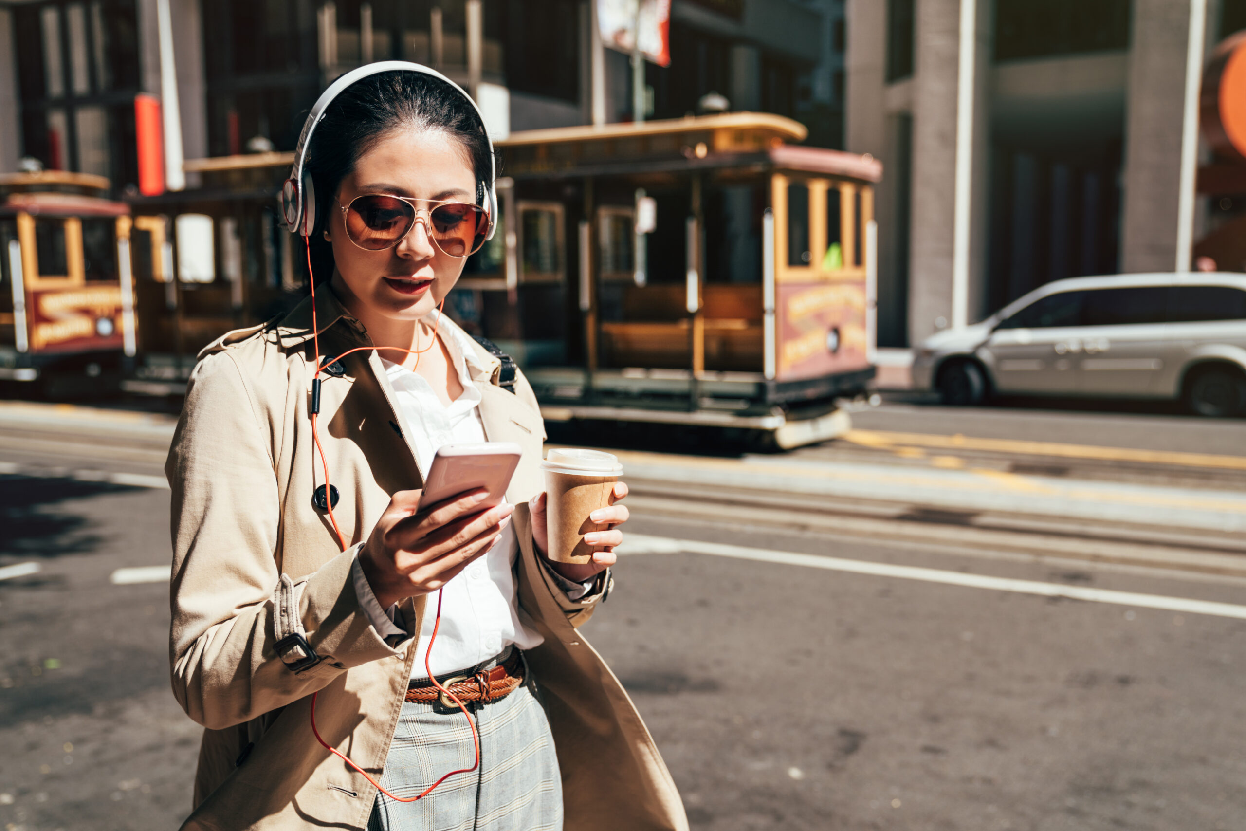 A woman walks on a street, wearing headphones, sunglasses, and holding a coffee cup and phone, with a cable car in the background.