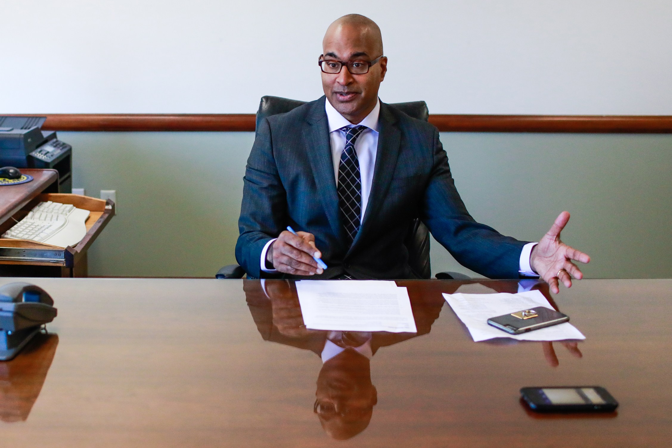 A person in a suit sits at a wooden desk with papers, a pen, and two phones. They're gesturing with one hand, looking engaged in conversation.