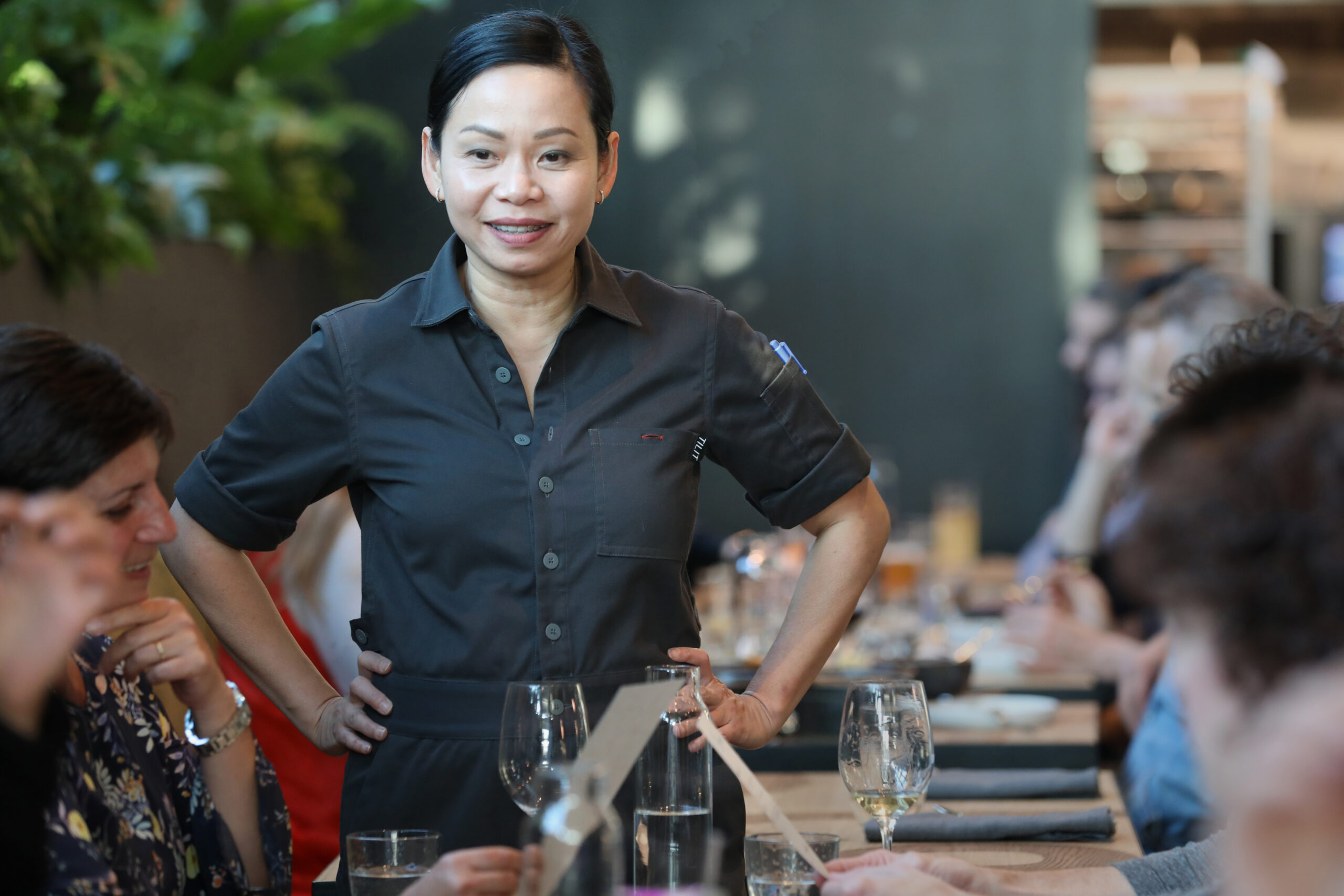 A woman in a gray uniform stands with hands on hips, smiling in a restaurant setting with people seated at a long table. There are plants and table settings visible.