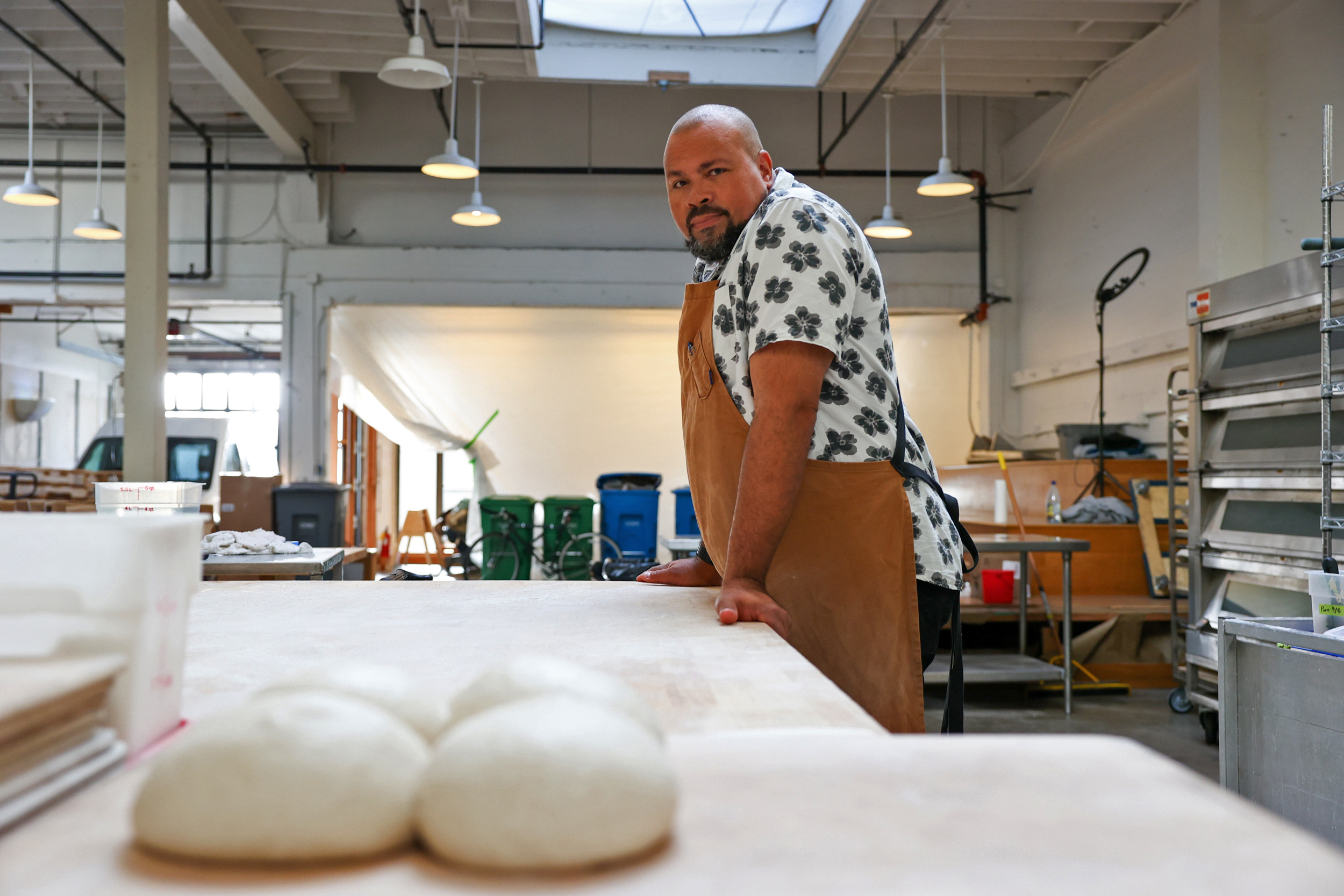 A shaven-headed man wearing a patterned short-sleeve shirt and a khaki apron stands next to a flat white table. Round balls of sourdough sit within his reach.