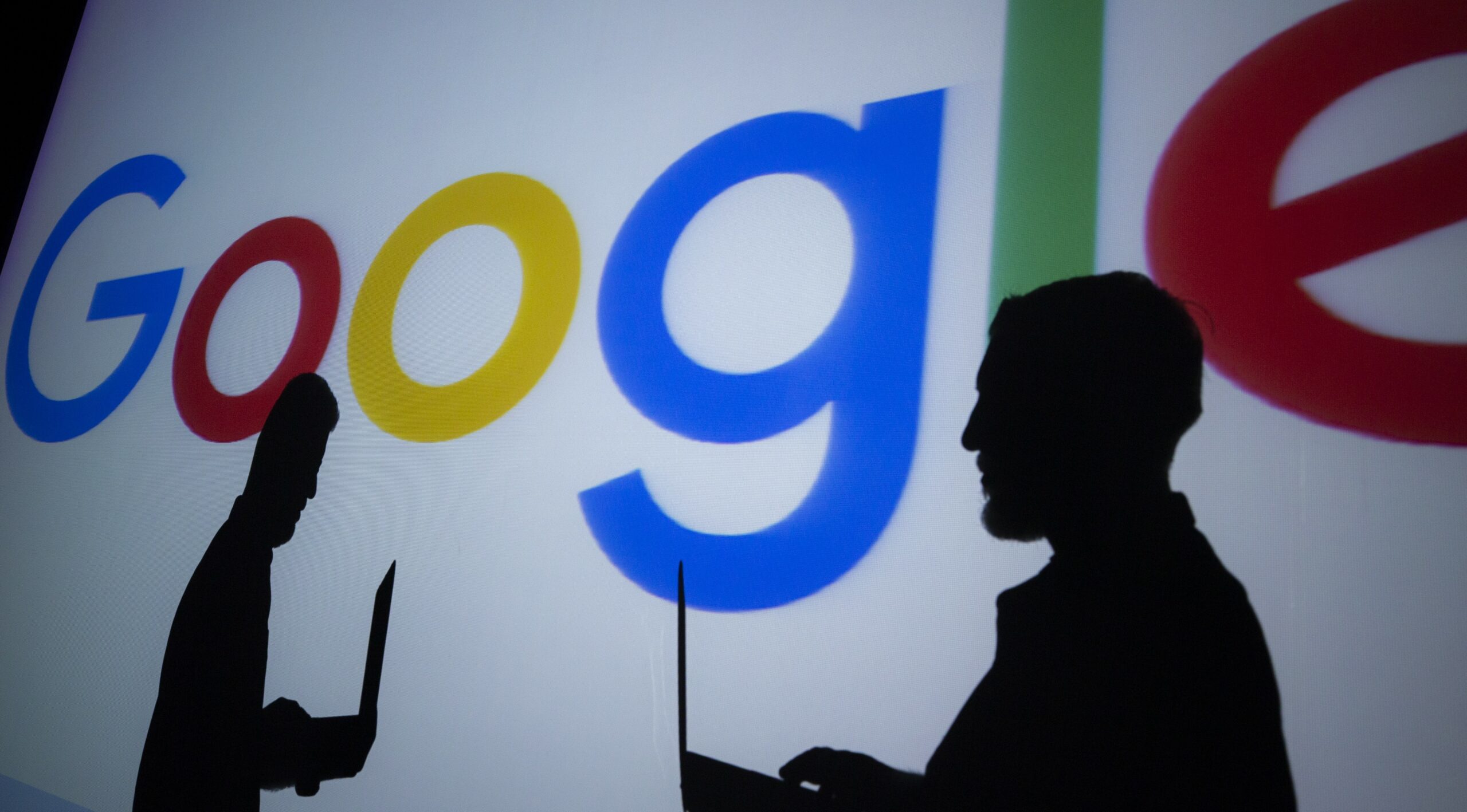 Silhouettes of people holding laptops are seen in front of the logo of 'Google' technology company on the 20th anniversary of Google.
