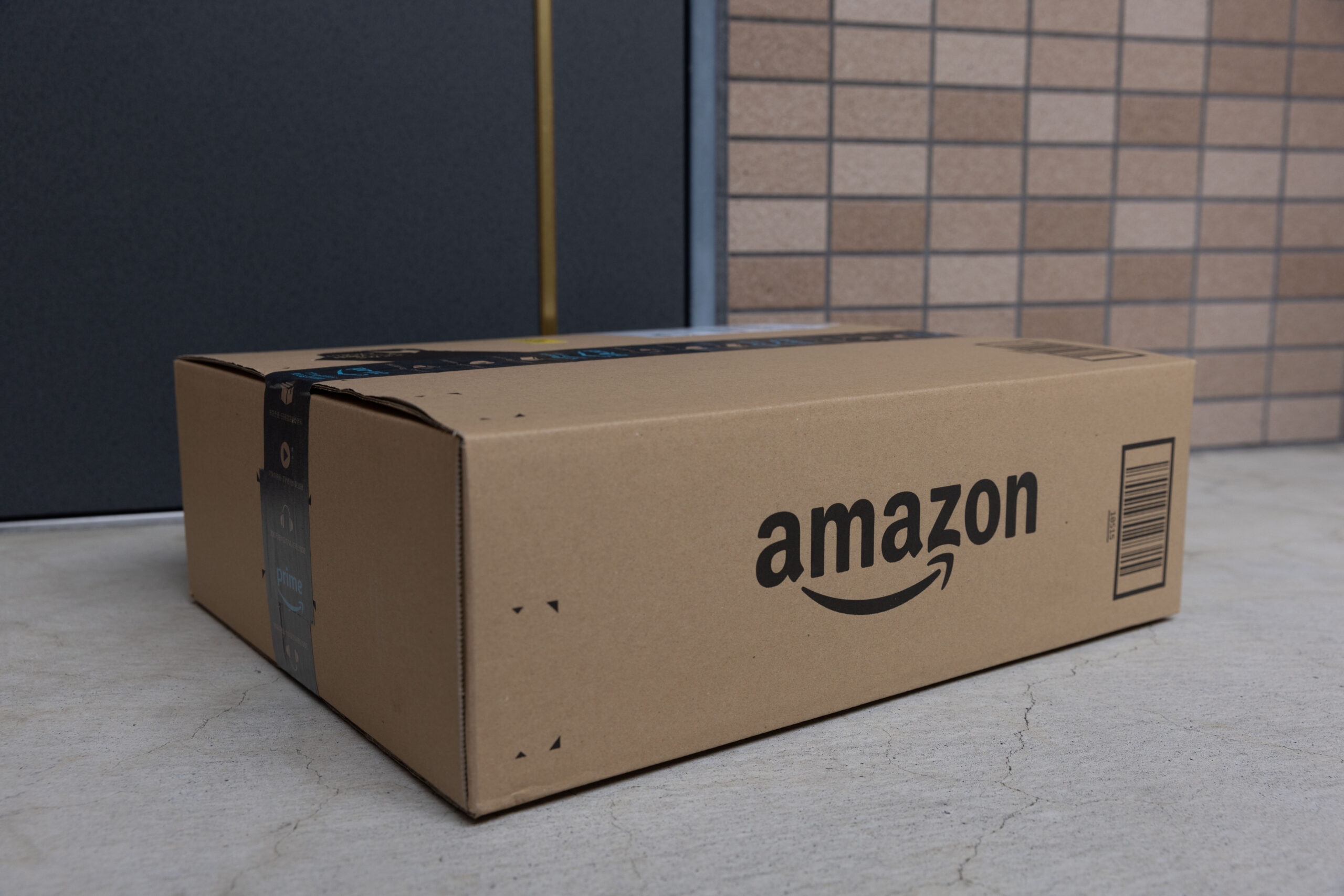 A box with the Amazon logo is on a gray doorstep. In the background, a blue door and a tiled wall.