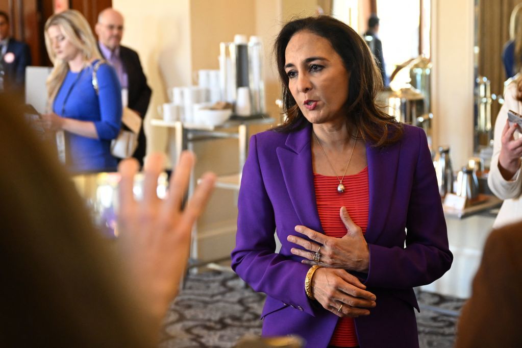 A woman in a purple blazer and red shirt is speaking to someone off-camera. She has a thoughtful expression and gestures with her hands. Others are in the background.