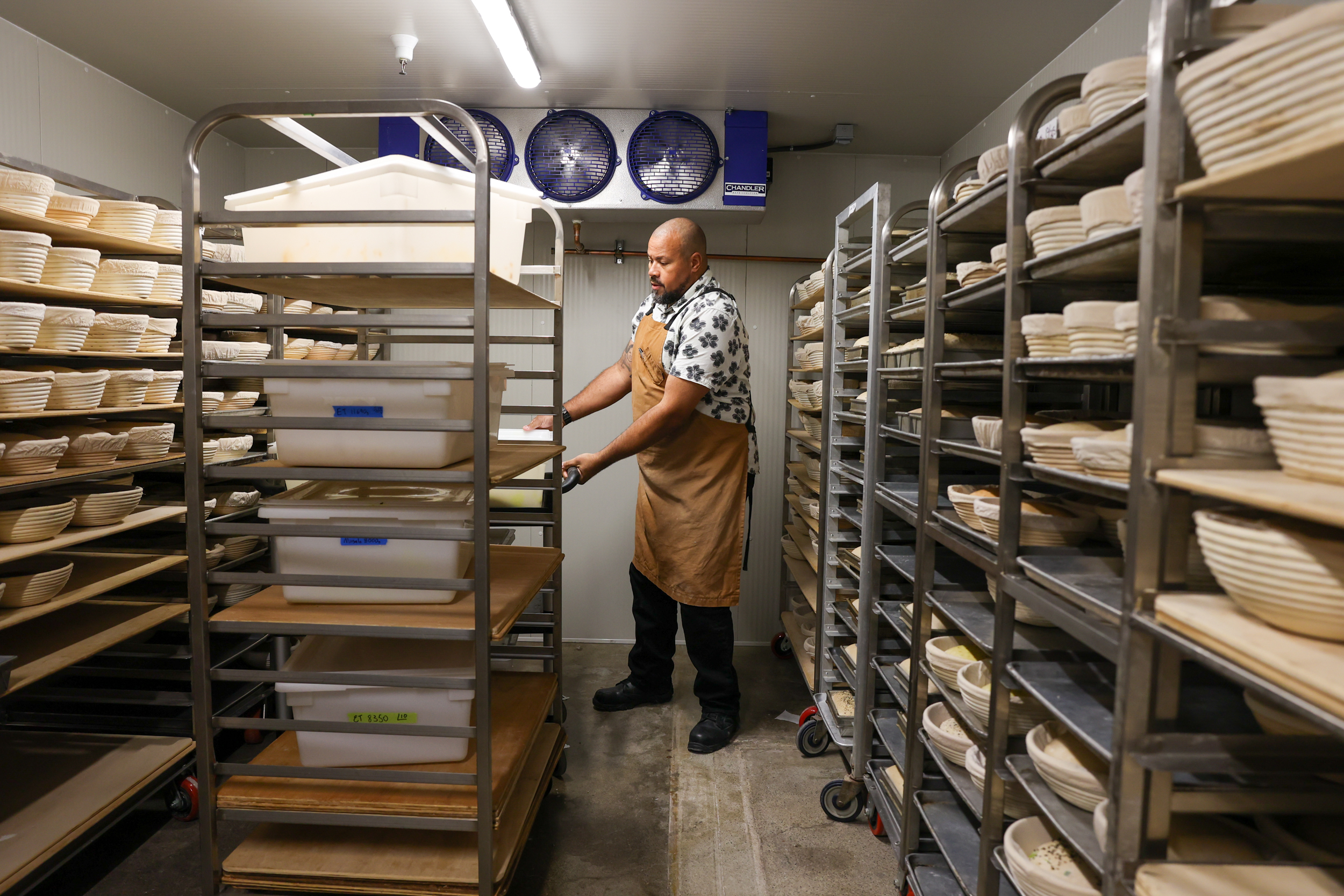 A shaven-headen man stands between tall metal shelf racks laden with dough and bread.