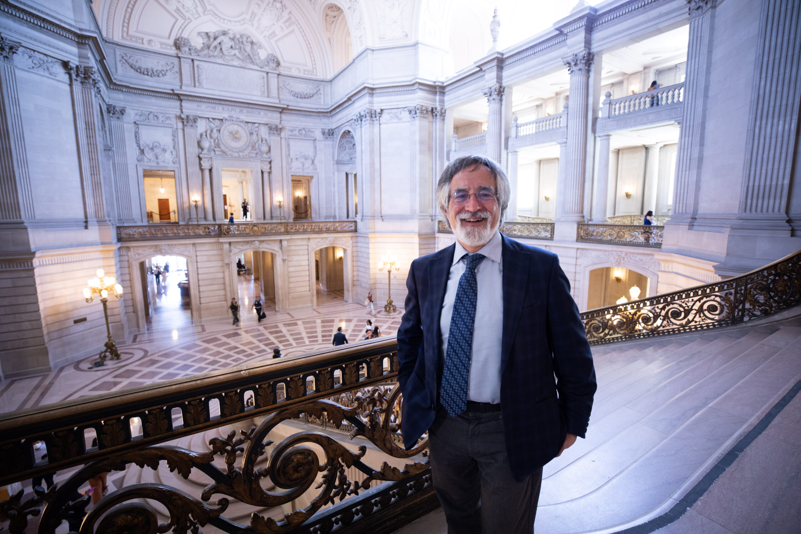 Supervisor Aaron Peskin in a suit stands on an ornate staircase in San Francisco's City Hall.