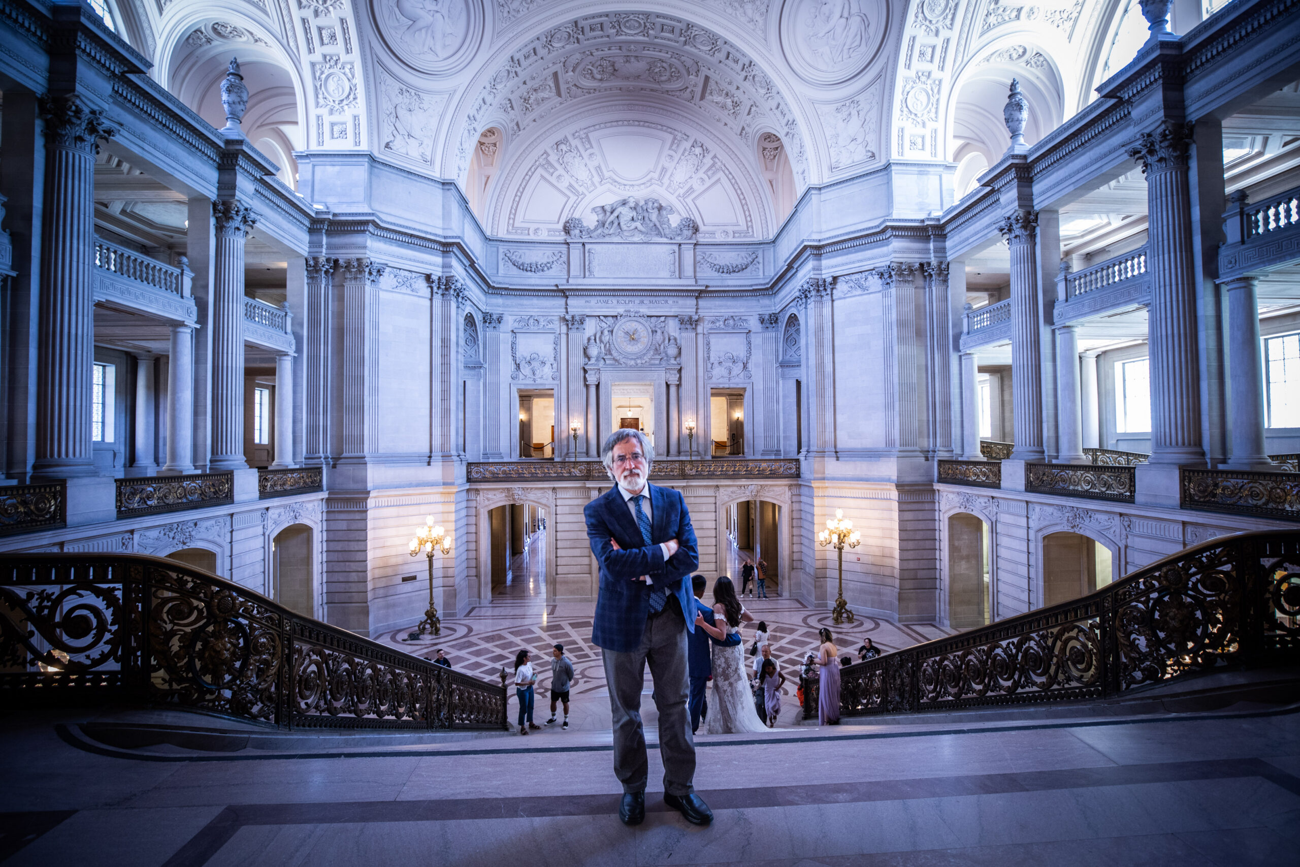 A man stands on an ornate staircase inside a grand, neoclassical hall with intricate architectural details.