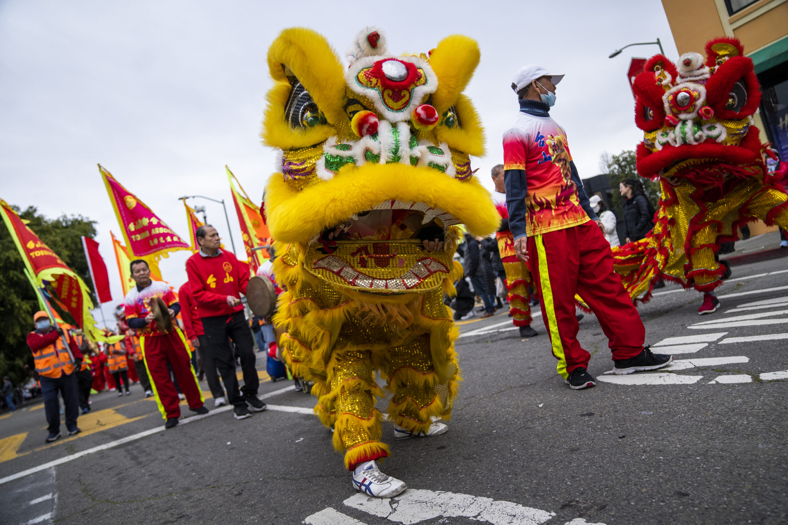 The image shows a vibrant street parade with performers in colorful lion costumes, lead by a bright yellow lion, accompanied by participants waving red and yellow flags.