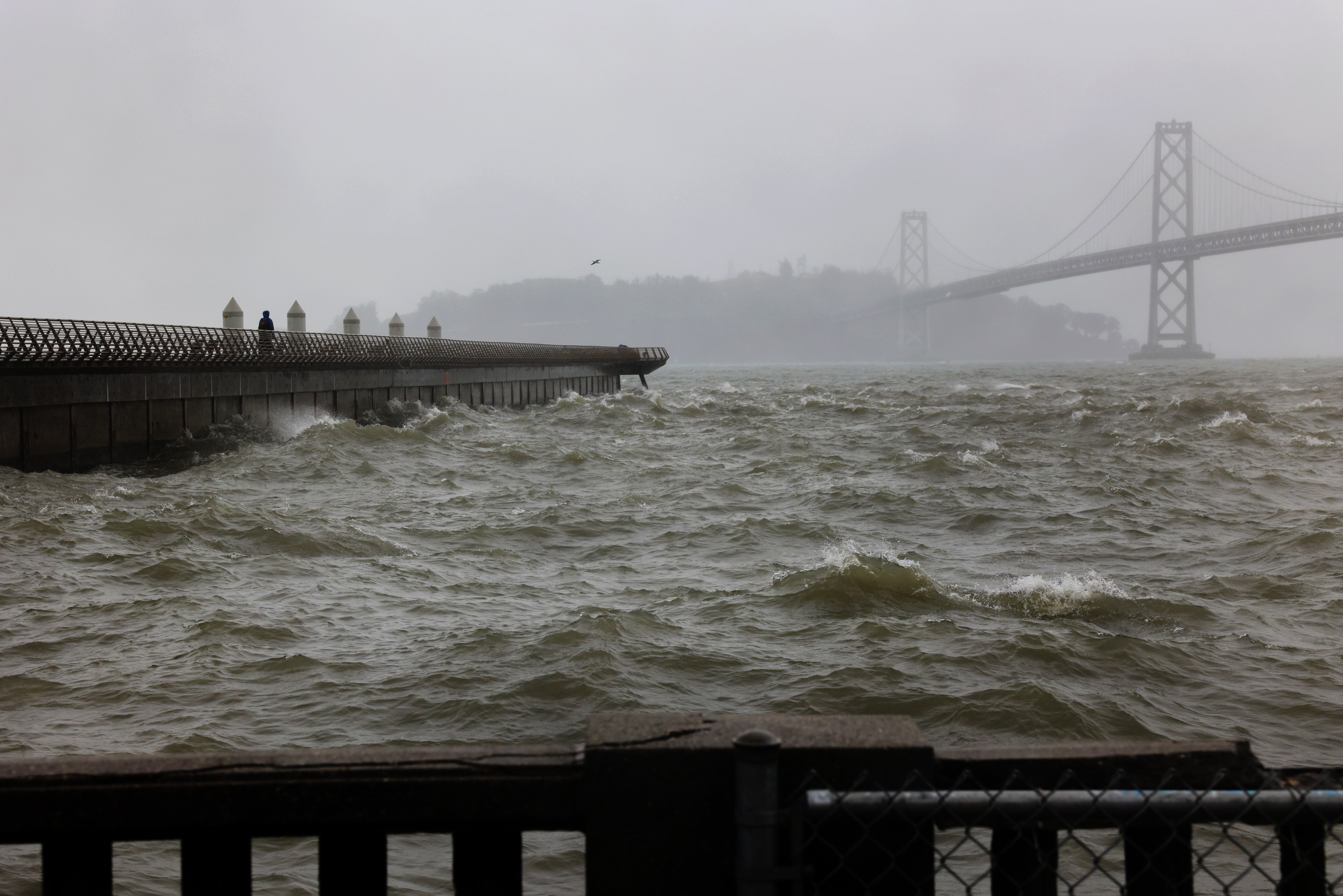 Choppy waters before a pier with a person; a bridge faintly looms in the misty background.