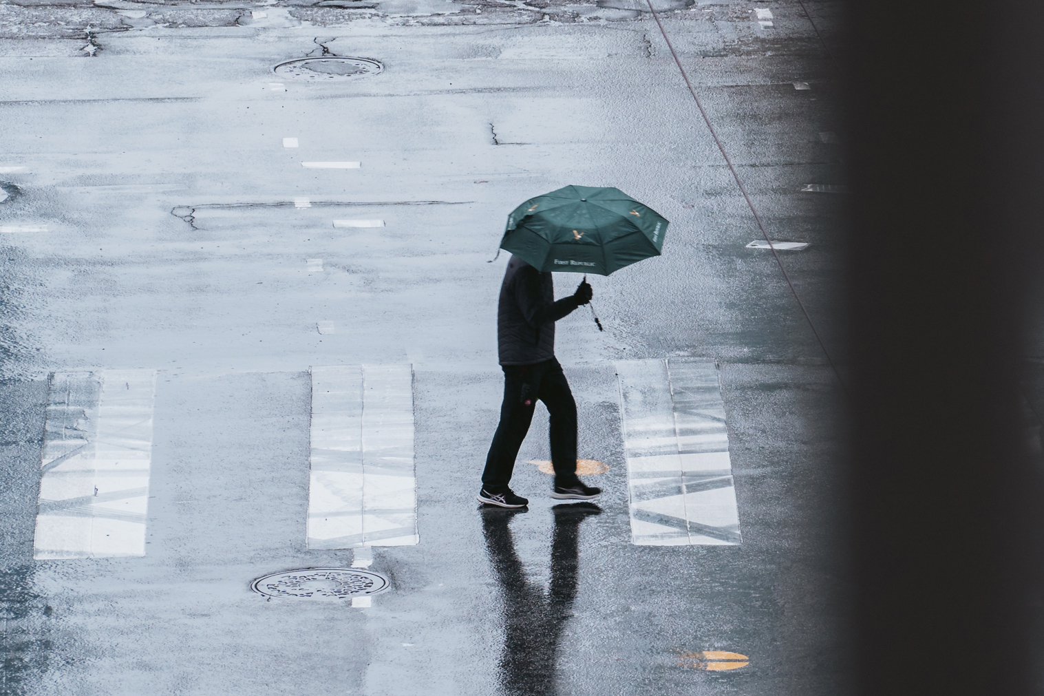 A distant, desaturated image of a pedestrian holding an umbrella while walking in a crosswalk