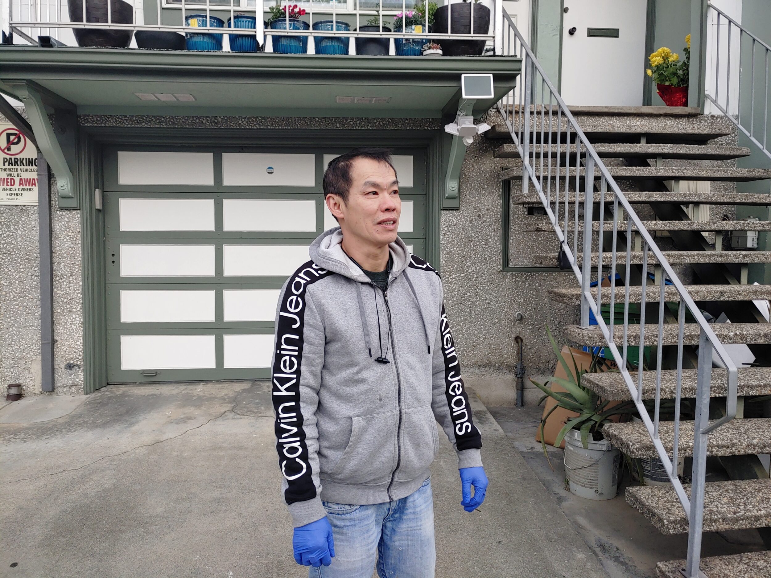 An Asian man in a Calvin Klein hoodie, jeans and blue nitrile gloves stands on a driveway in front of a green and white garage door.