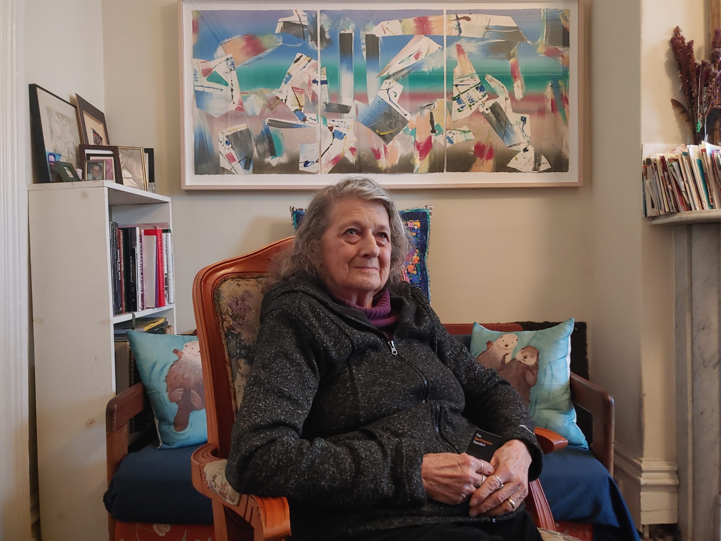 An older woman sits in a vintage chair with an abstract artwork in the background.