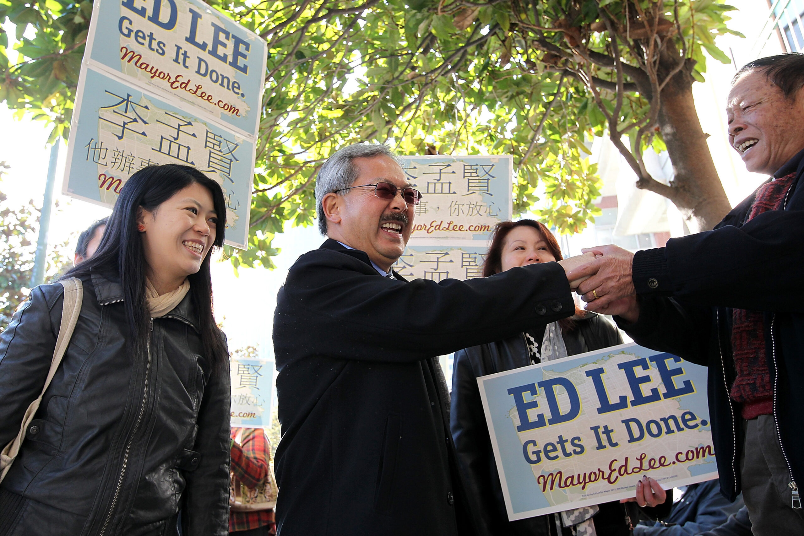 The late Mayor Ed Lee, a man with gray hair wearing sunglasses, is seen from the waist up shaking someone's hands. Supporters hold &quot;Ed Lee&quot; signs.