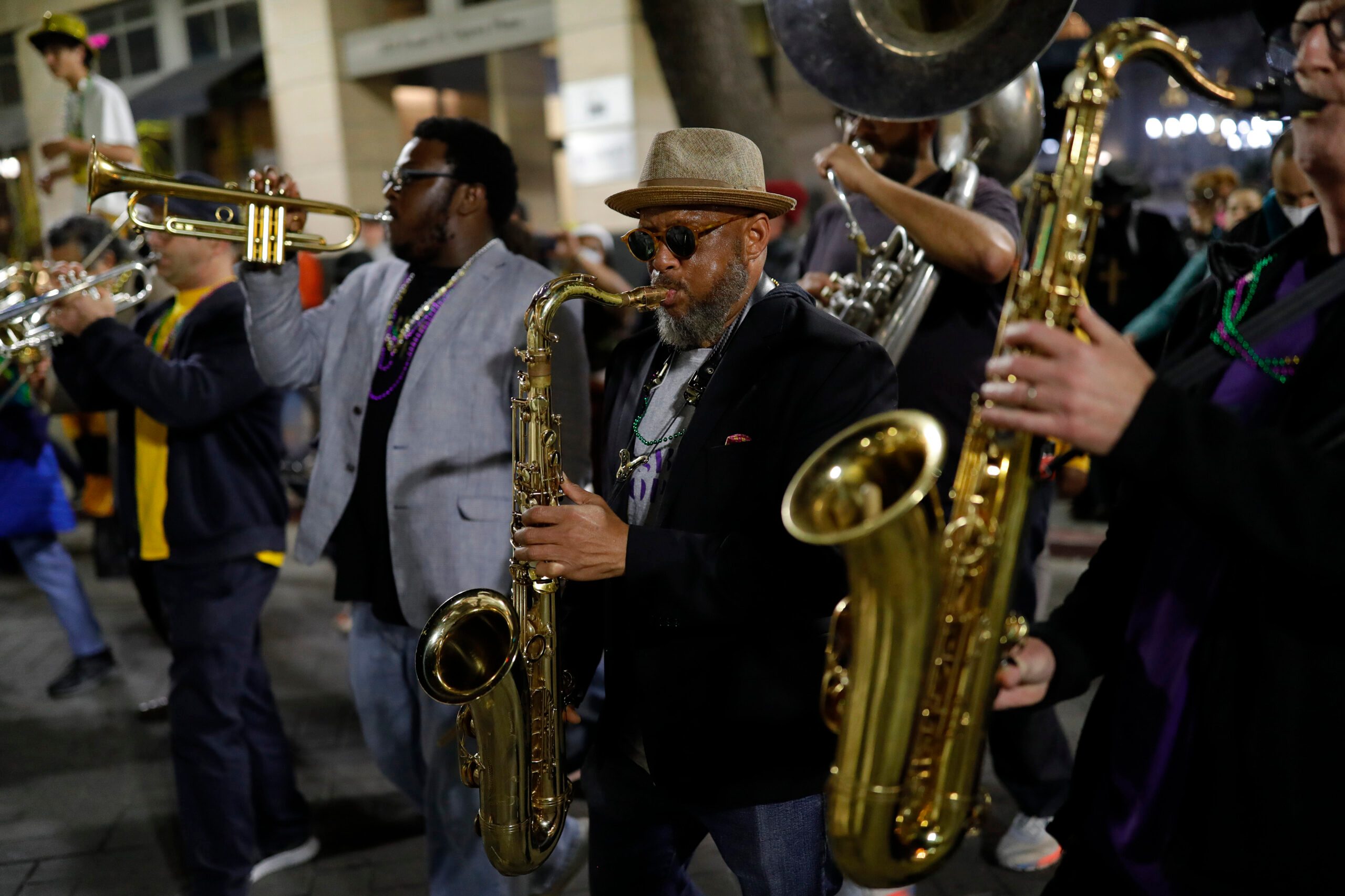 A lively brass band plays in a parade, with musicians wearing beads. They play trumpets, saxophones, and other instruments, dressed casually with hats and sunglasses.