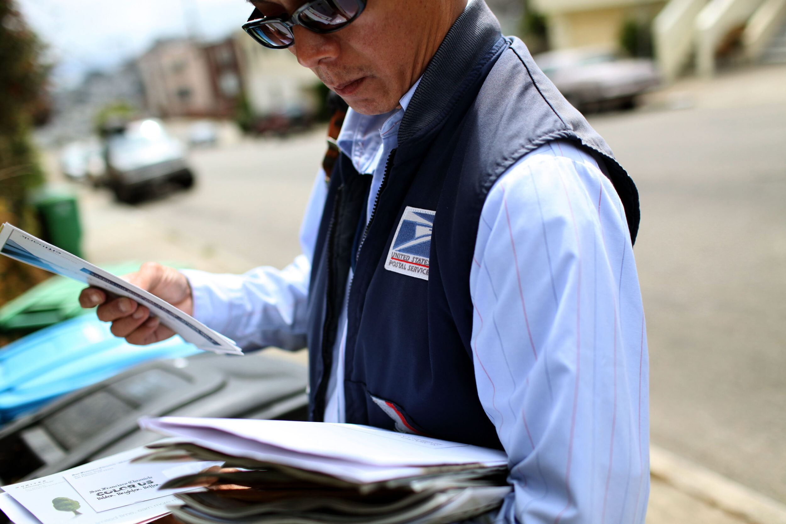 A postal worker is sorting mail on a street, wearing sunglasses and a vest.