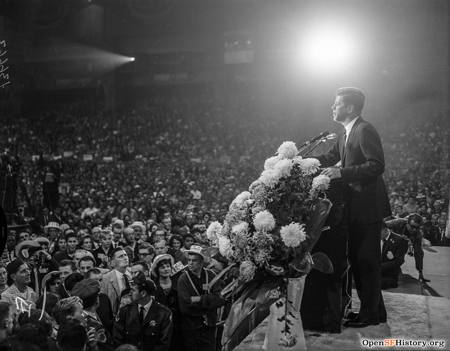 President John F. Kennedy speaks at a podium with flowers, facing a large, attentive crowd in an arena.