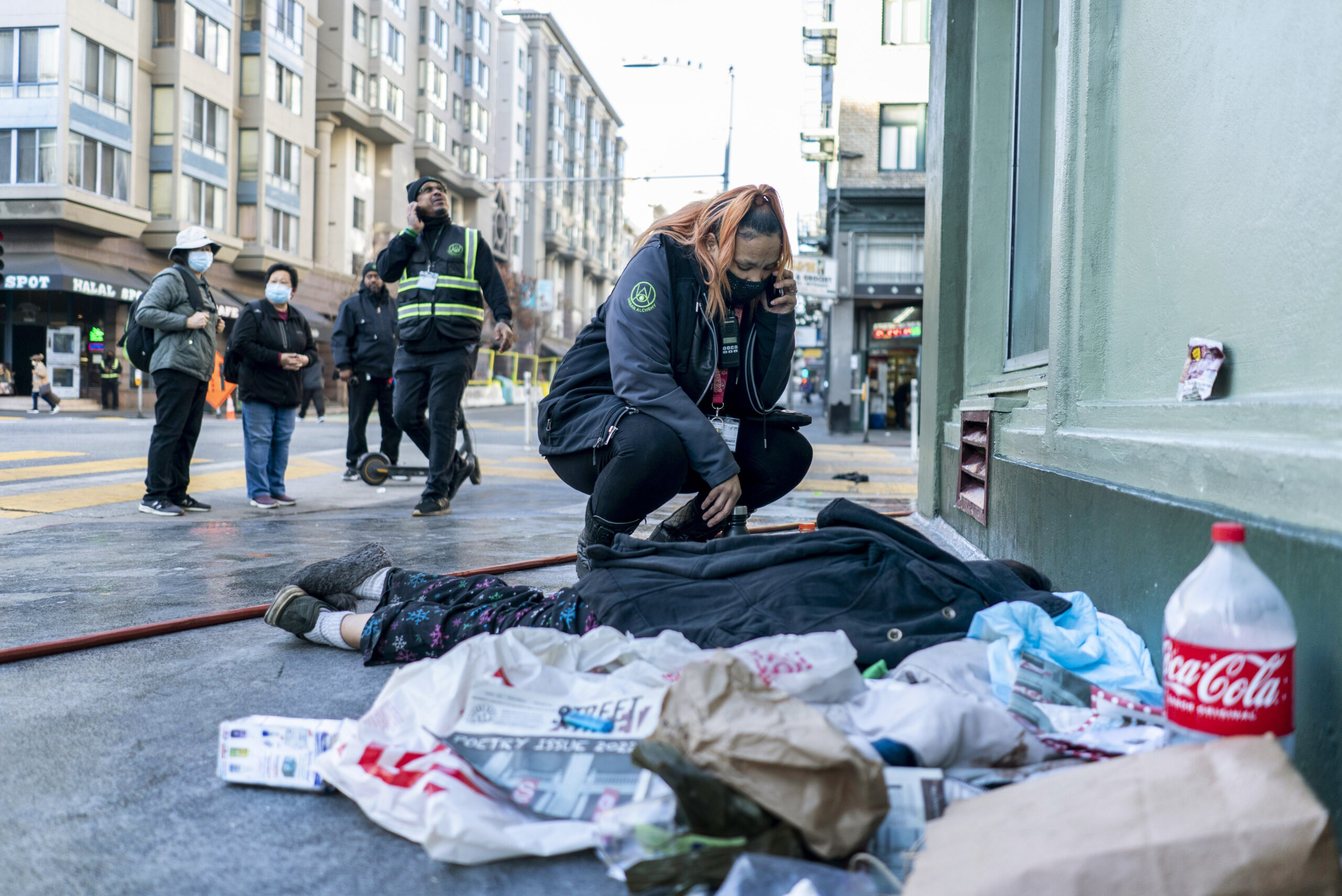 In an urban setting, a woman crouches and speaks on a phone next to a person lying on the ground. Onlookers stand nearby amidst scattered belongings and a Coca-Cola bottle.