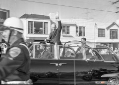 President Dwight Eisenhower waves from a car in a parade, flanked by motorcyclists, with onlookers in the background.
