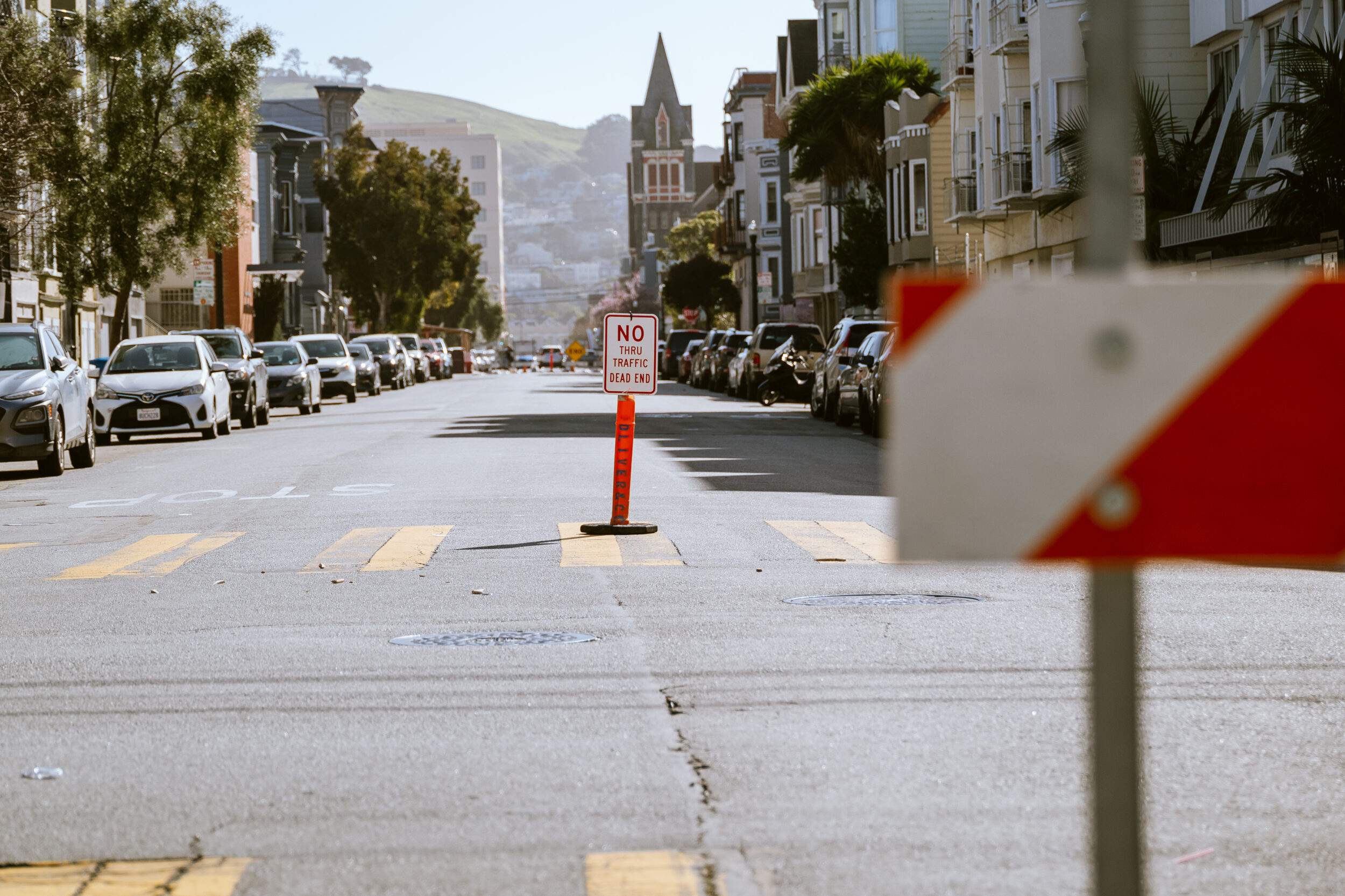 A traffic sign sits on an orange delineator on a San Francisco street on a sunny day with hills in the background.