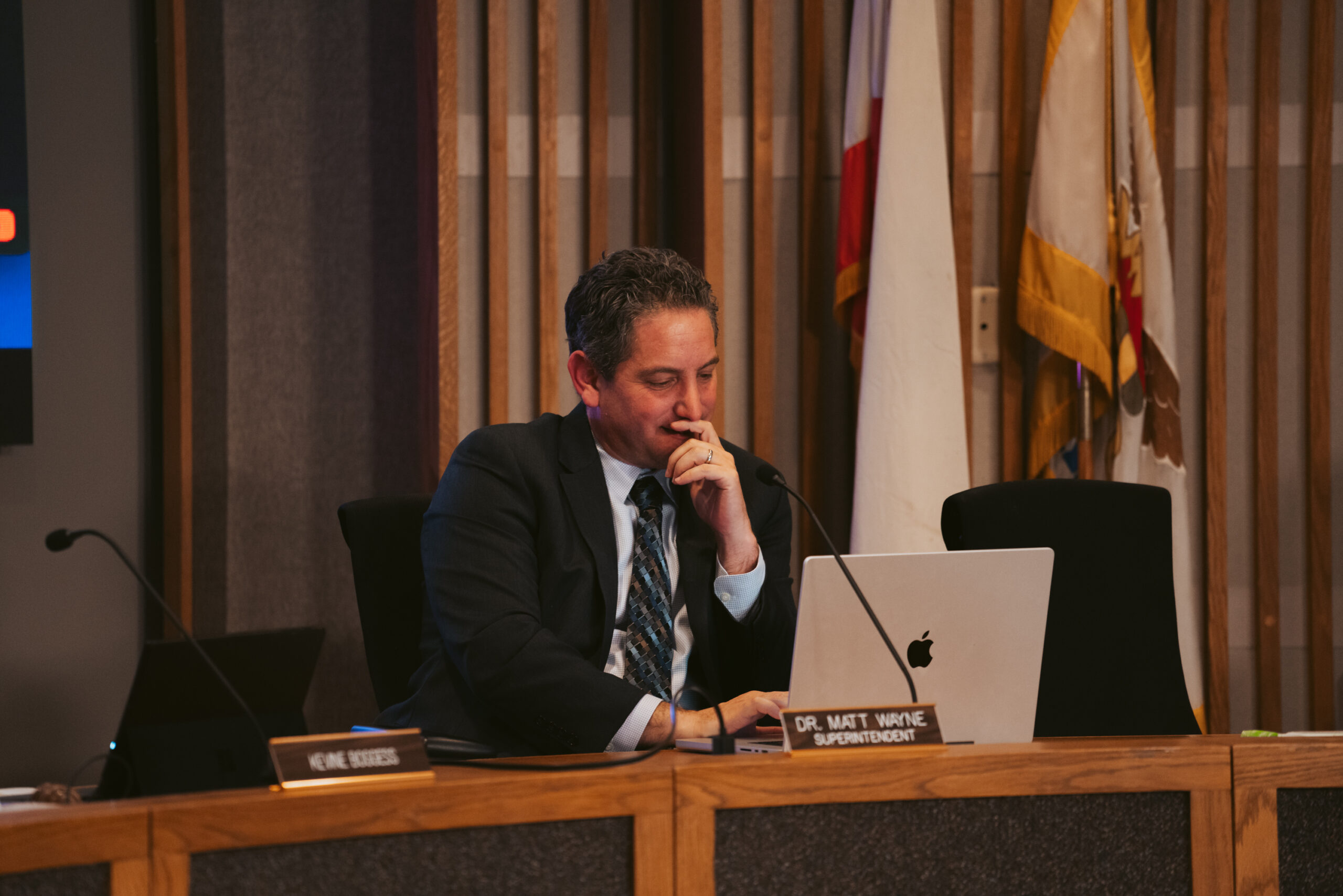 A man sits at a desk looking at a laptop, deep in thought. He's wearing a suit, and in the background are flags. The nameplate reads &quot;Dr. Matt Wayne, Superintendent.&quot;