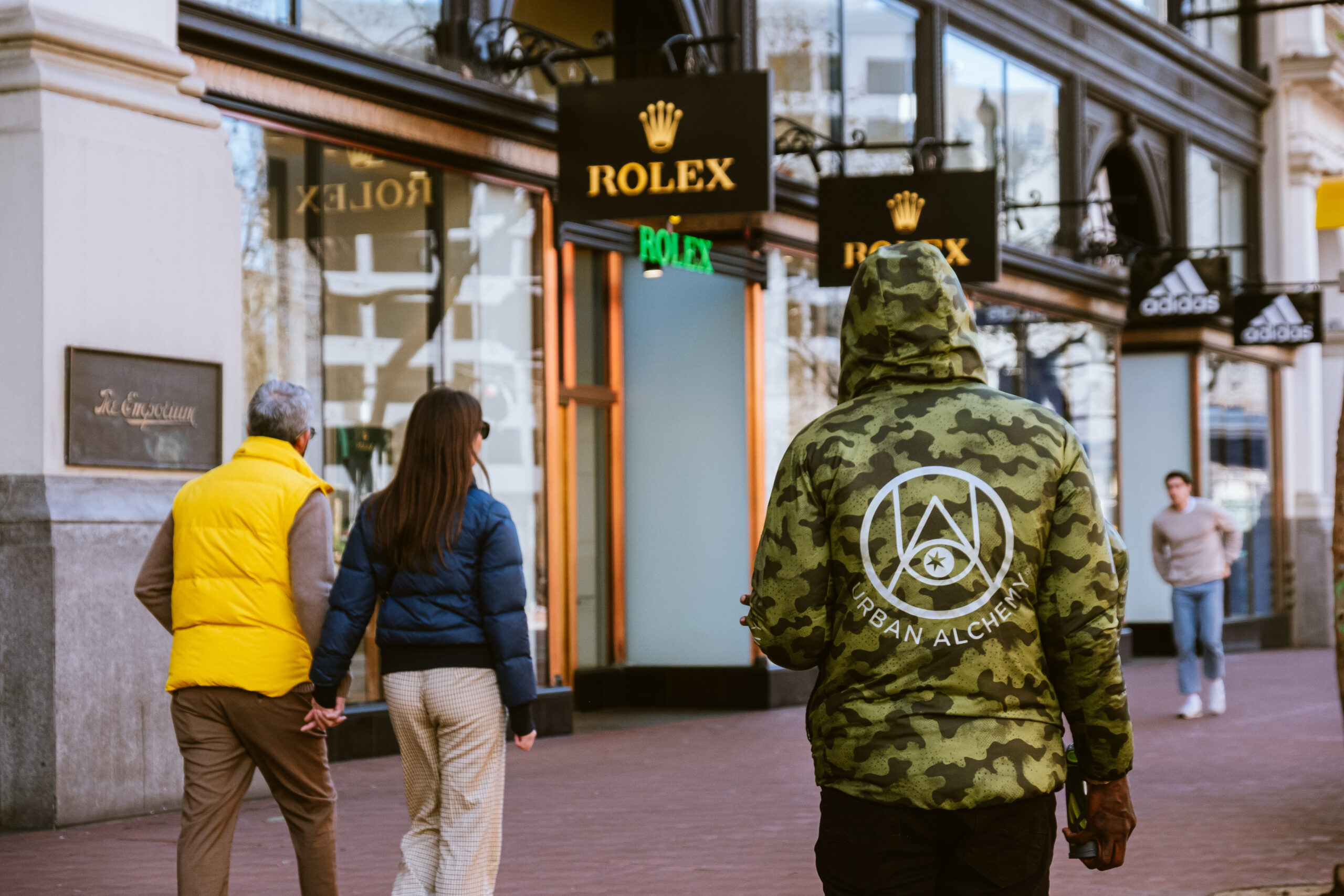 A person in a camo hoodie walks past Rolex stores; a couple holding hands is also visible.