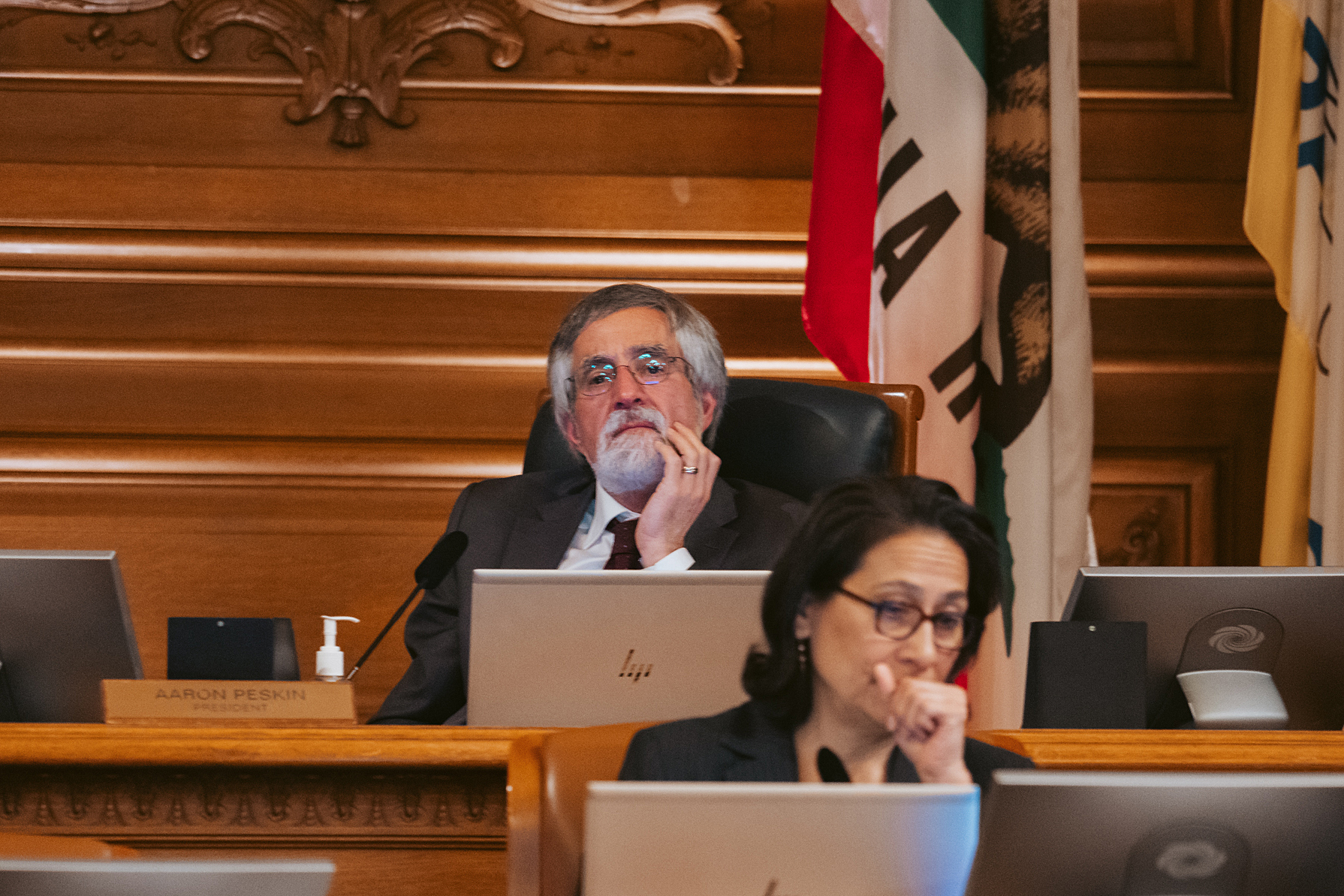 Aaron Peskin sits contemplatively in the San Francisco Board of Supervisors chambers with a hand on his chin. A woman, also absorbed, is partially visible in the foreground.
