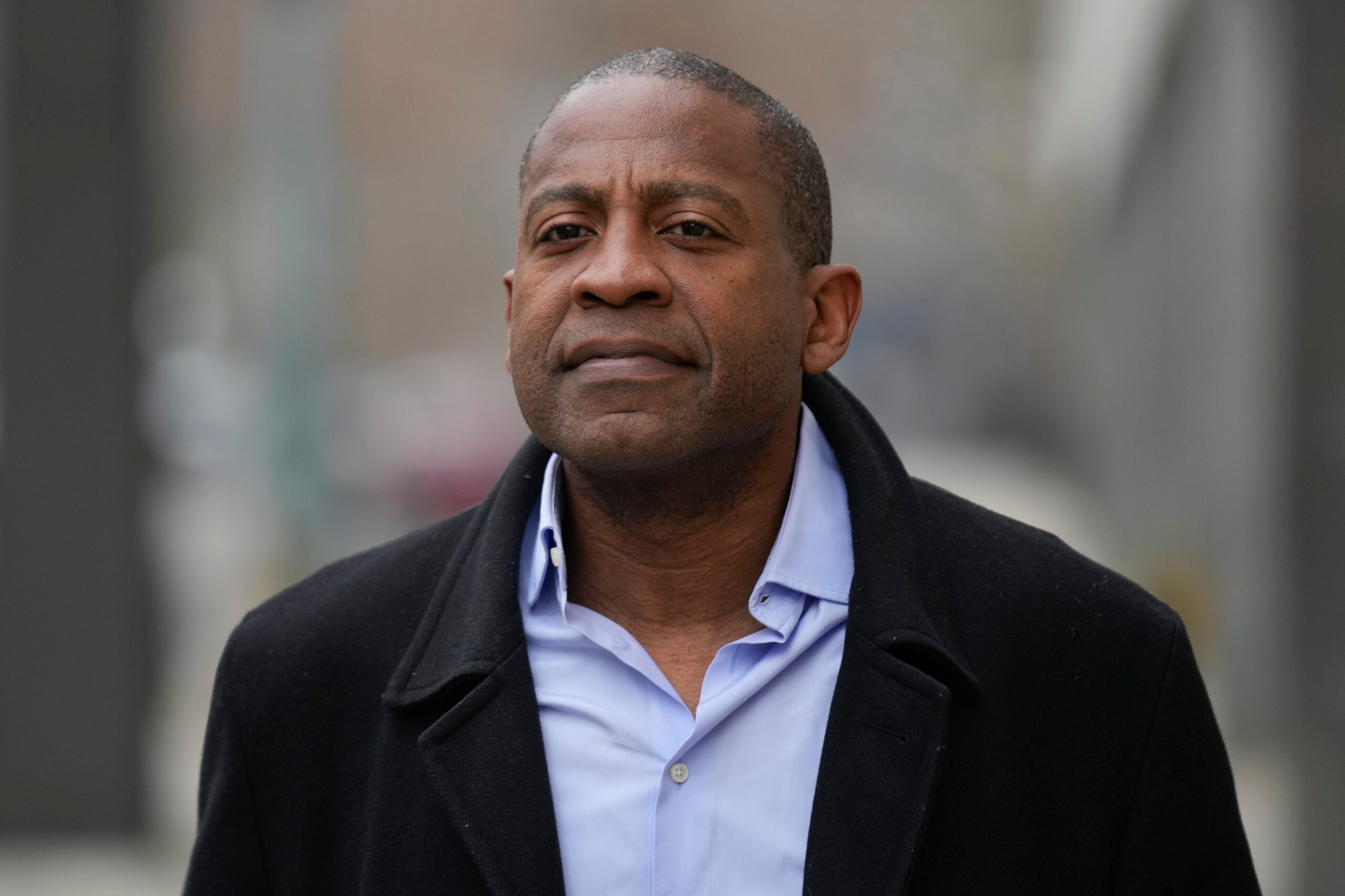 A man is walking outside wearing a dark overcoat and a light blue shirt. The background is blurred, suggesting an urban environment.