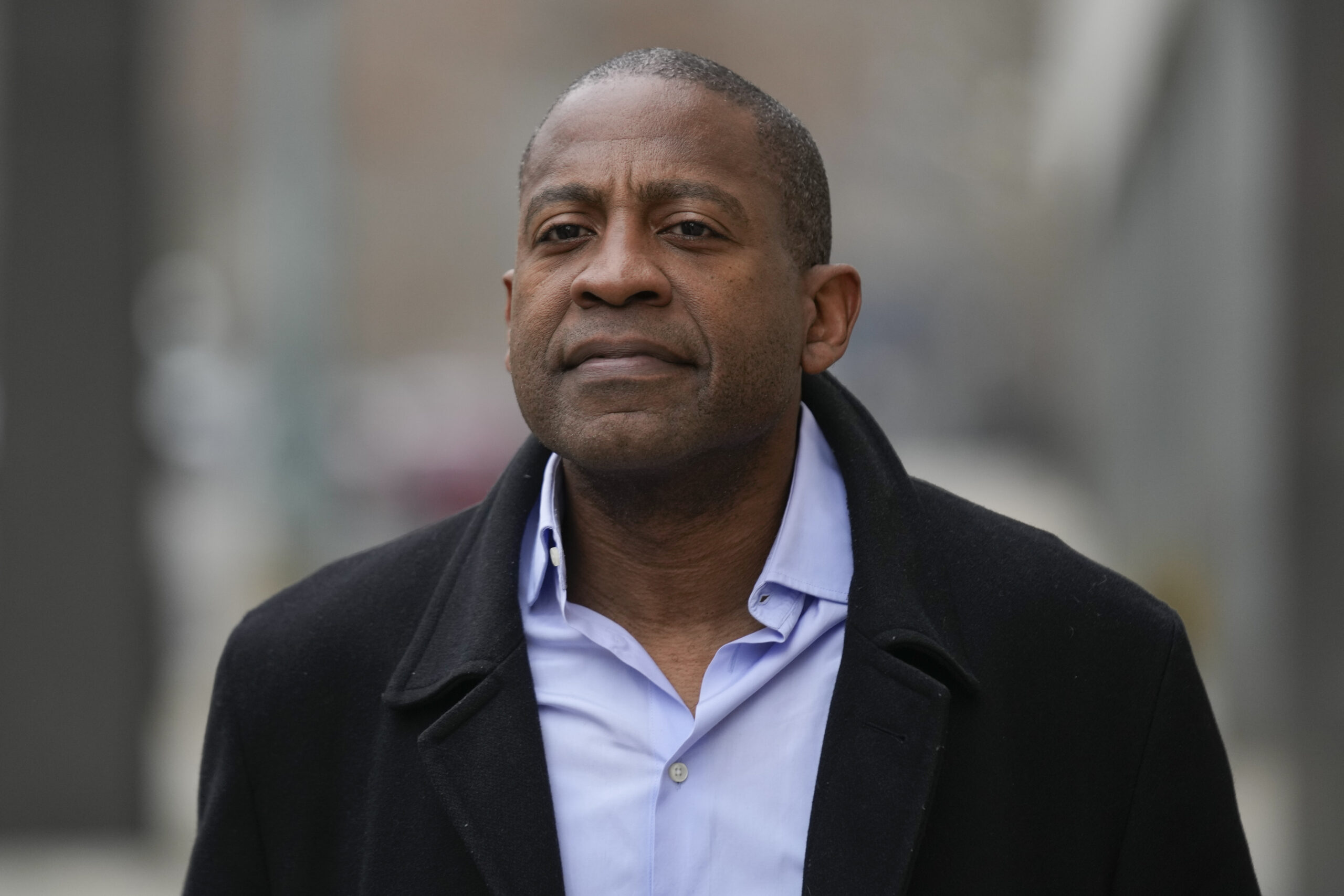 A man is walking outside wearing a dark overcoat and a light blue shirt. The background is blurred, suggesting an urban environment.