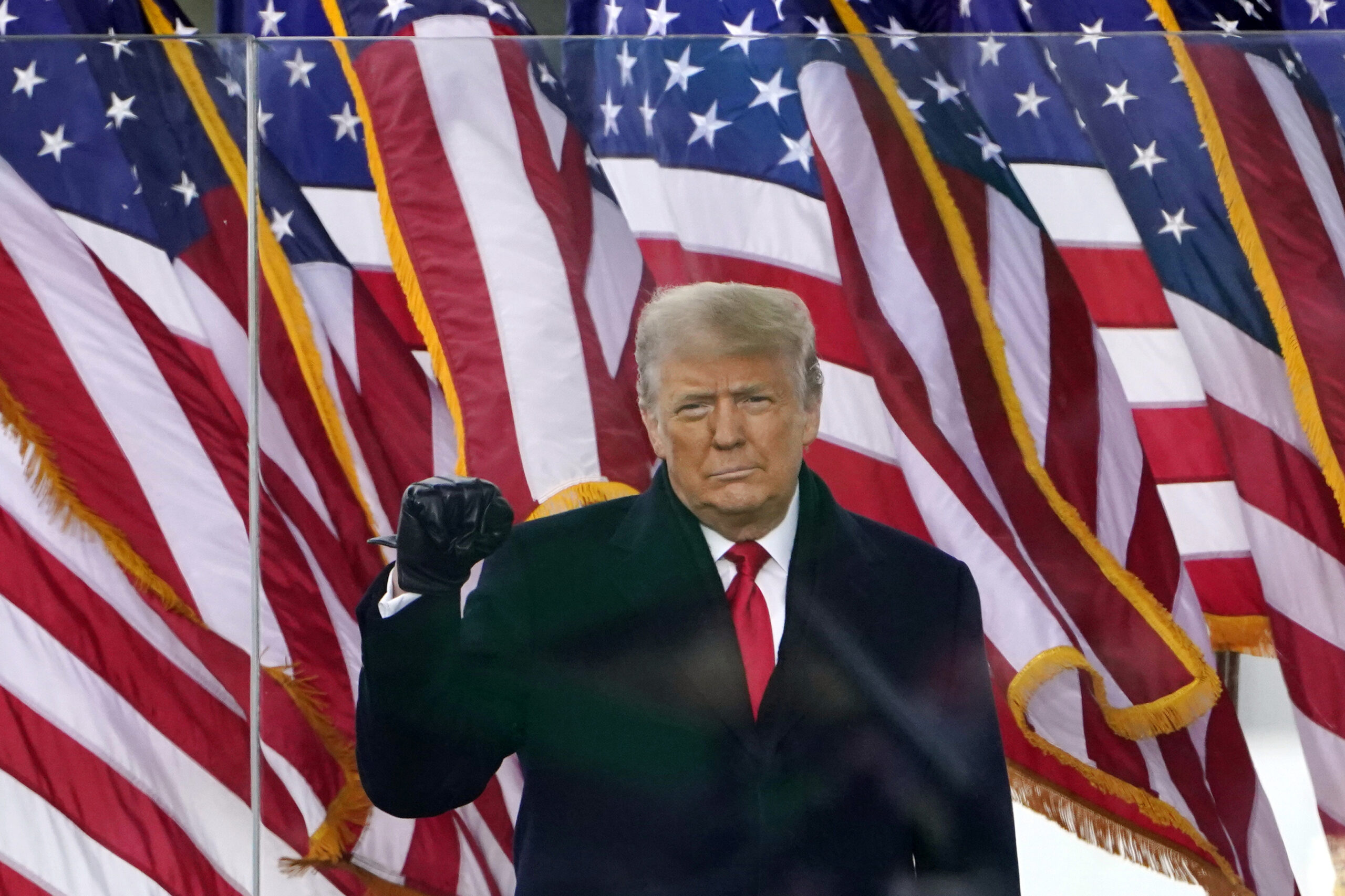 Donald Trump stands in front of multiple American flags, wearing a dark suit, white shirt, and red tie.