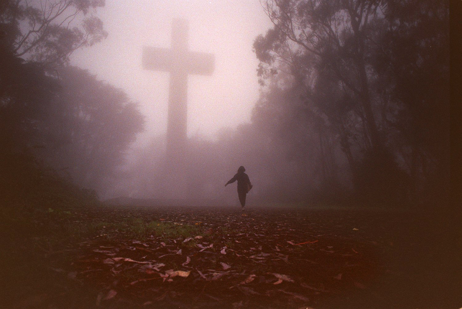 A silhouetted person walks towards a large cross in a foggy, forested area with a ground covered in leaves.