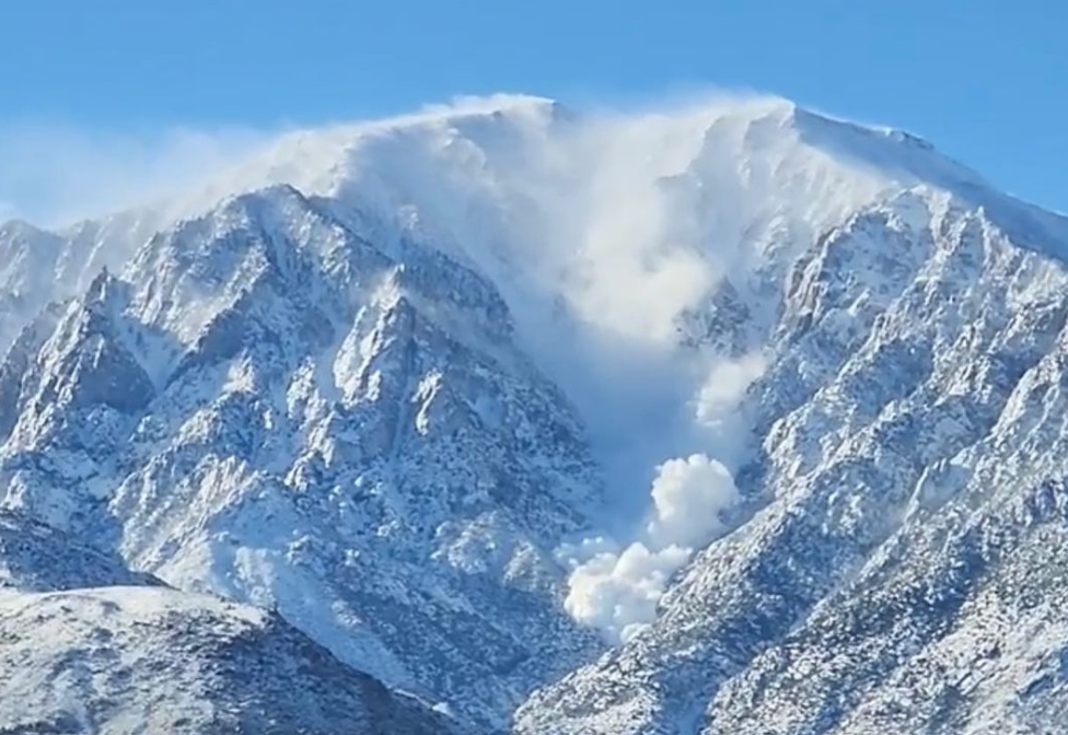 Snow-covered mountain peak with wind blowing snow off the top under a clear blue sky.