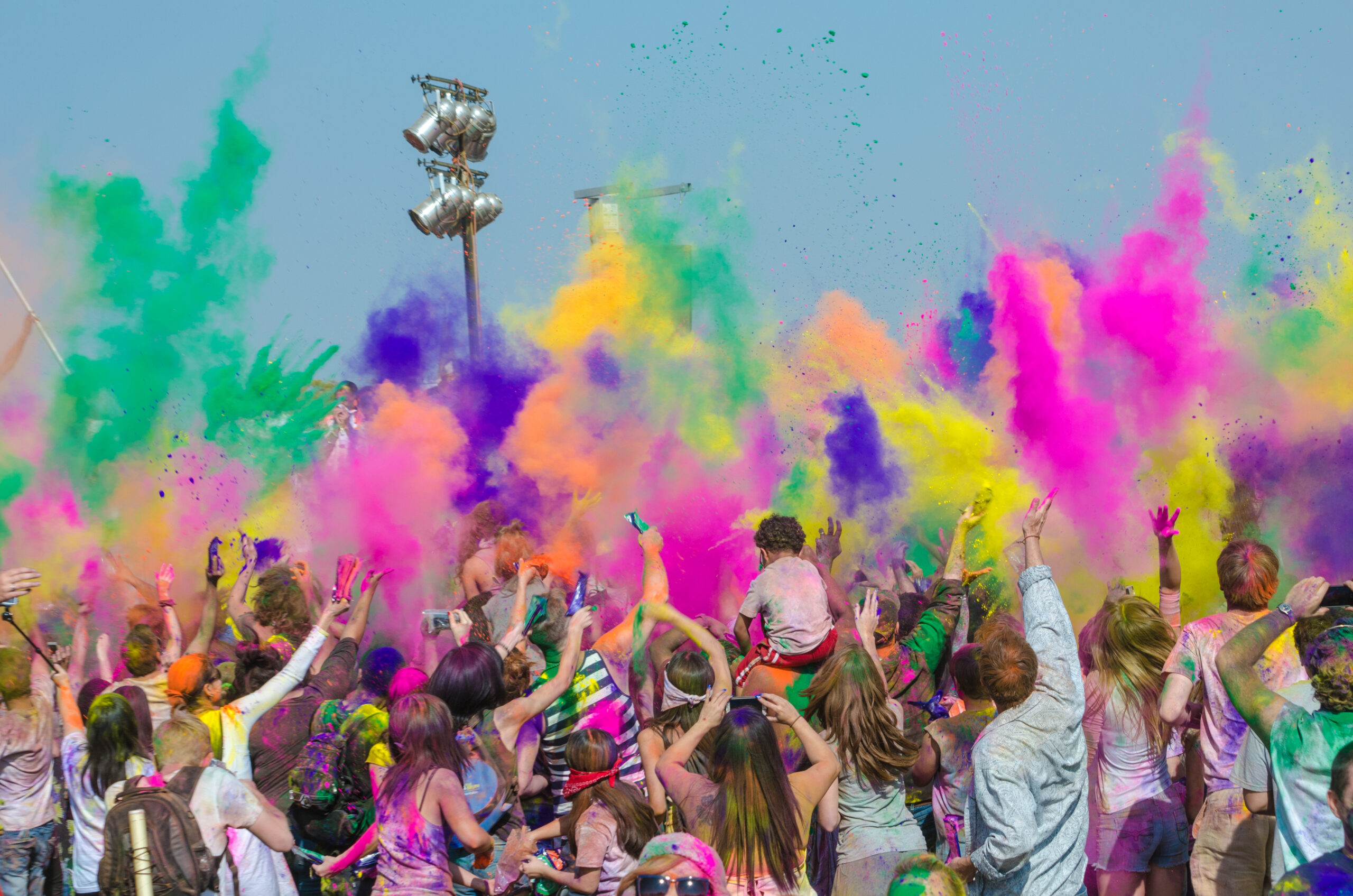 A jubilant crowd celebrates by tossing vibrant colored powders into the air, creating a rainbow explosion against a clear blue sky.