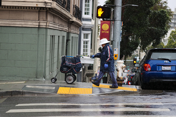 A man in a US Postal Service uniform walks on a wet sidewalk